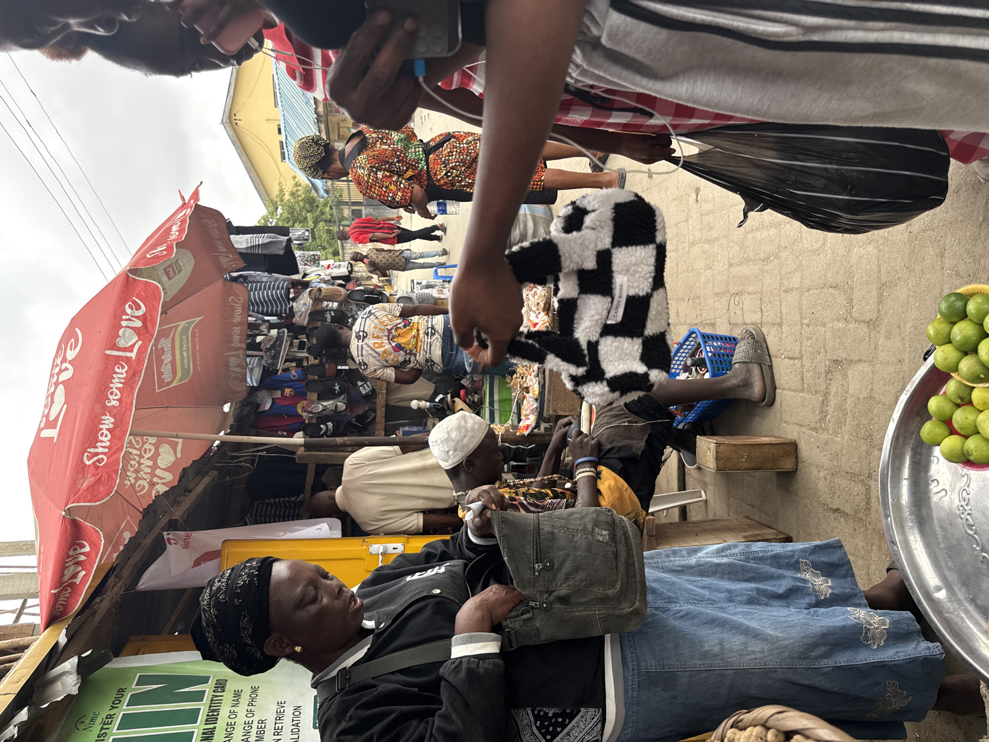 In Lekki, Nigeria, a street scene unfolds under a red umbrella displaying the text "Show some Love". A woman in a black headscarf and jacket, with a denim skirt, is reaching into a dark grey shoulder bag. Beside her, a man wearing a white skullcap and a yellow and brown patterned garment is seated, working with what appears to be beads or jewelry. In the foreground, a metal tray holds a collection of green limes. Further in the background, various stalls display clothing and footwear. Other individuals are visible, including a person in a vibrant, patterned dress, and others walking through the market. The ground is paved with concrete blocks.