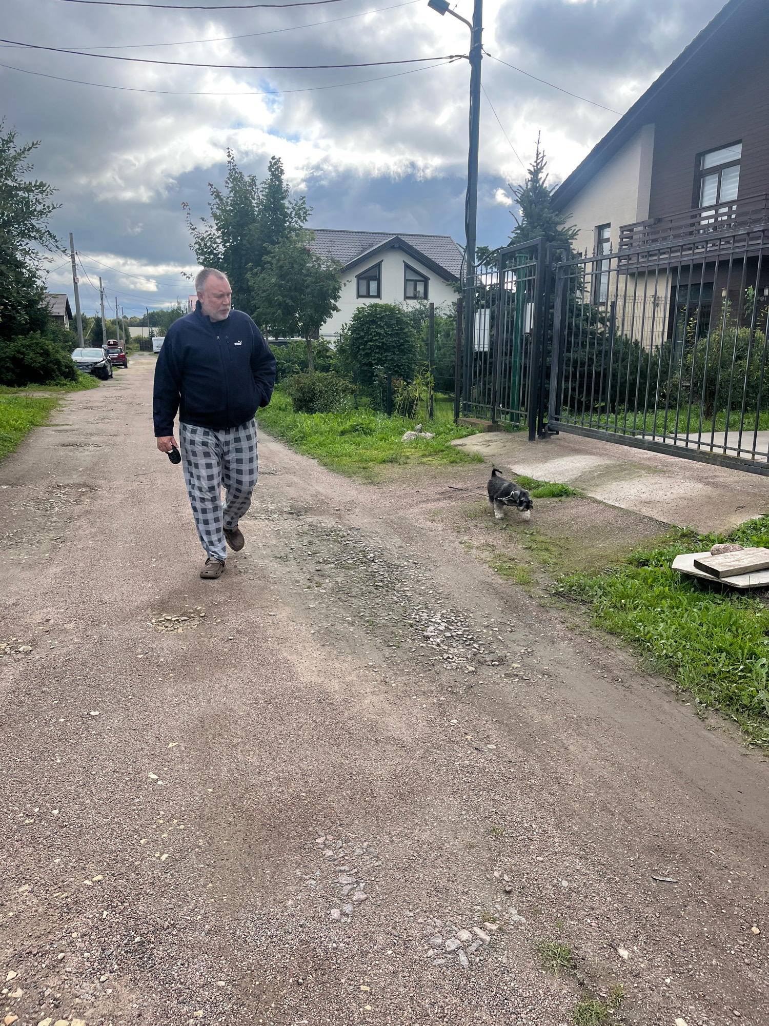An older white male with a beard walks down a gravel road in Vladimirovka, Russia. He is wearing a dark blue jacket and plaid pants, and he is holding a black object in his left hand. A small black and tan dog on a leash walks to his right, a few feet ahead of him. The road is lined with green grass and trees. In the background, there are houses and a black metal gate. The sky is cloudy.