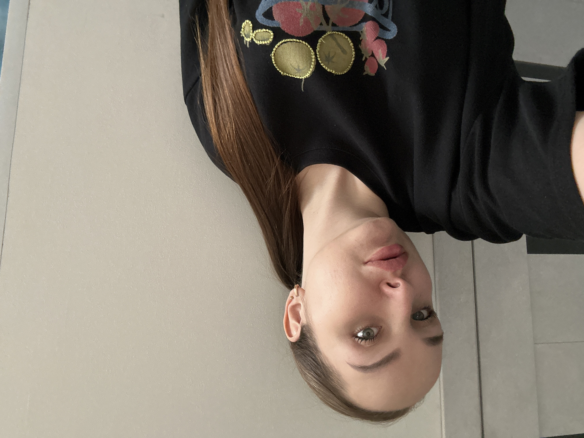 A selfie photograph shows a woman with fair skin, light brown hair, and green eyes. She is wearing a black sweatshirt with a planet and fruit design. The background appears to be a plain wall, possibly in an indoor setting. There is no indication of the location being in Chaikovsky, Russia, or any specific city.
