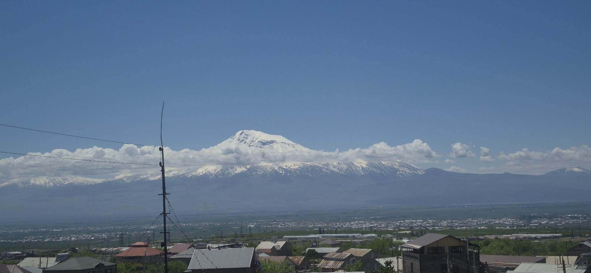 The image shows a wide view of a landscape with a mountain range in the background, likely taken from an elevated position within a city or town. The mountain range is partially covered with snow and surrounded by clouds. Below the mountains, there's a densely populated area with numerous buildings, likely houses, and some greenery interspersed. In the foreground, rooftops of several buildings are visible, providing a sense of depth to the image. A utility pole with wires extending across the frame is situated on the left side of the image, slightly obscuring the view. The sky is a clear, bright blue, indicating a sunny day.