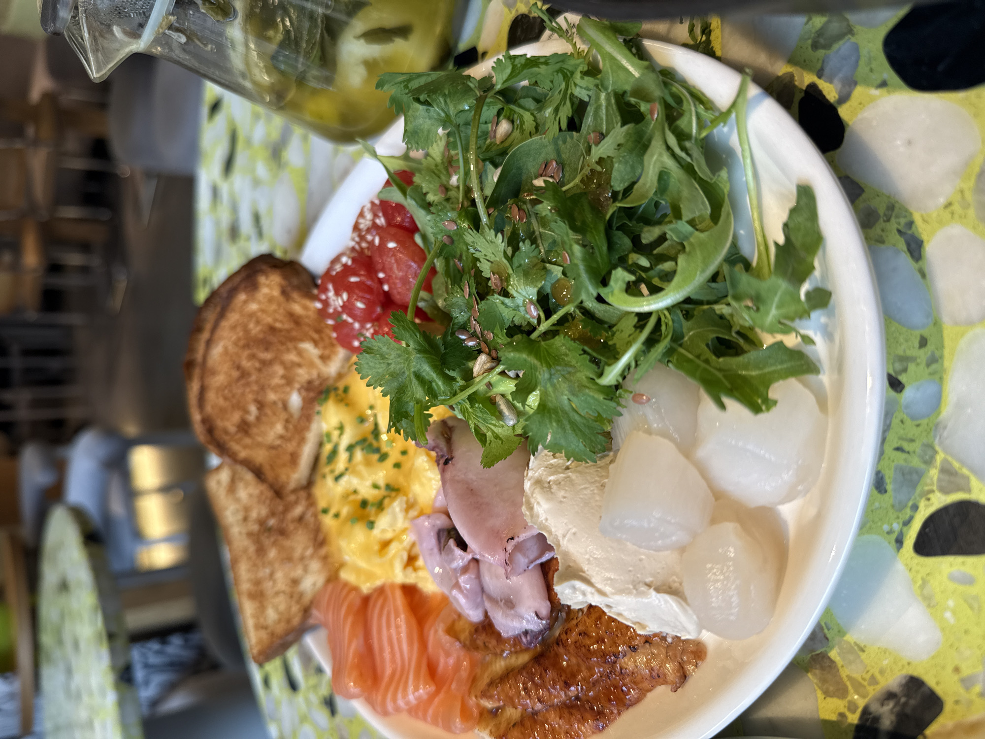 A close-up, overhead shot shows a white plate filled with a breakfast or brunch spread, placed on a table with a bright yellow and green terrazzo pattern. To the left of the plate are two slices of toasted bread. Next to the toast, moving clockwise, are thinly sliced pieces of smoked salmon, a portion of scrambled eggs sprinkled with chives, and a pile of halved cherry tomatoes topped with sesame seeds. Further clockwise is a bed of leafy greens, likely arugula, dressed with oil and scattered with seeds. In the center of the plate, beneath the eggs and tomatoes, is a dollop of a creamy white spread, possibly hummus or a cheese spread, and several pieces of what appear to be pickled onions or lychee fruits. To the far right of the plate, a glass carafe containing a green liquid, possibly olive oil or infused water with herbs, is visible. The background is blurred, showing the interior of a restaurant with tables and chairs, suggesting an urban dining environment. The image is well-lit, highlighting the textures and colors of the food. No people are visible in the frame. The table's distinctive terrazzo pattern could potentially help identify the specific establishment in Saint Petersburg, Russia, if this pattern is unique to a particular restaurant or cafe in the city.