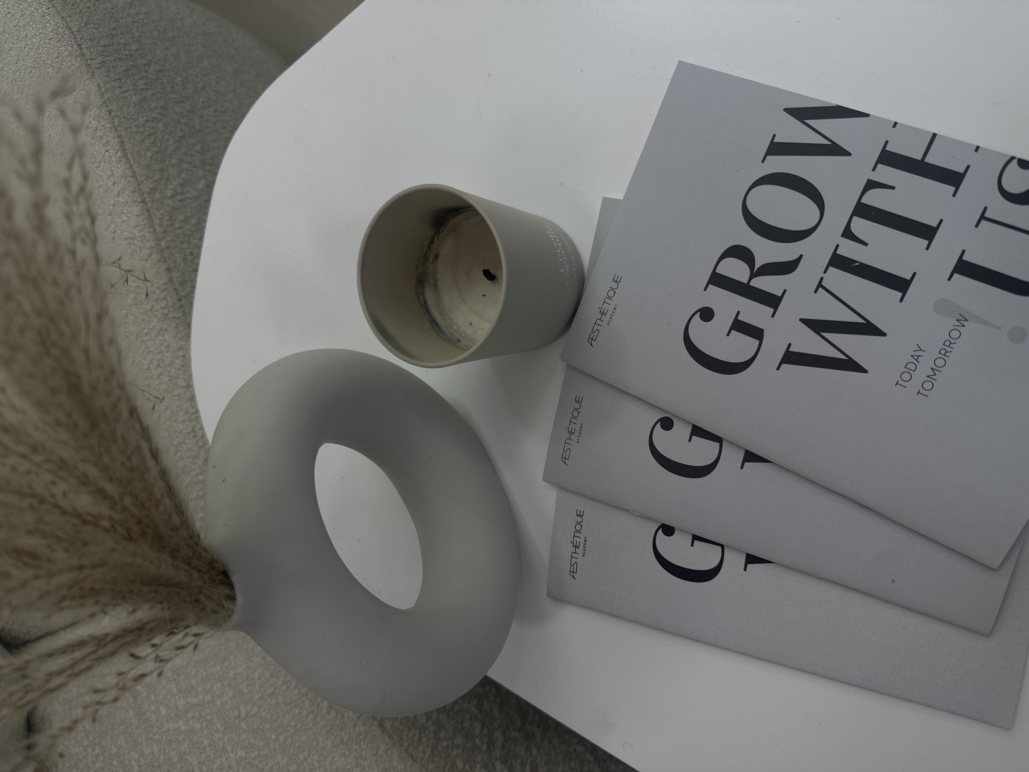 The image shows a tabletop scene, likely indoors, with a minimalist aesthetic. A donut-shaped matte gray vase containing dried pampas grass rests on the left. Beside the vase is a used gray candle holder, indicating a burned candle. To the right, a stack of gray leaflets are partially visible, with the words "GROW WITH US" printed in large, black capital letters on the topmost leaflet, and the words "AESTHÉTIQUE ACADEMY" printed in smaller font. The scene is set on a round, white tabletop, and part of a gray textured surface (likely a sofa or chair) is visible in the background. There's no explicit indication of Yerevan, Armenia, or any people or actions within the frame. The scene suggests a neutral, modern decorative style.