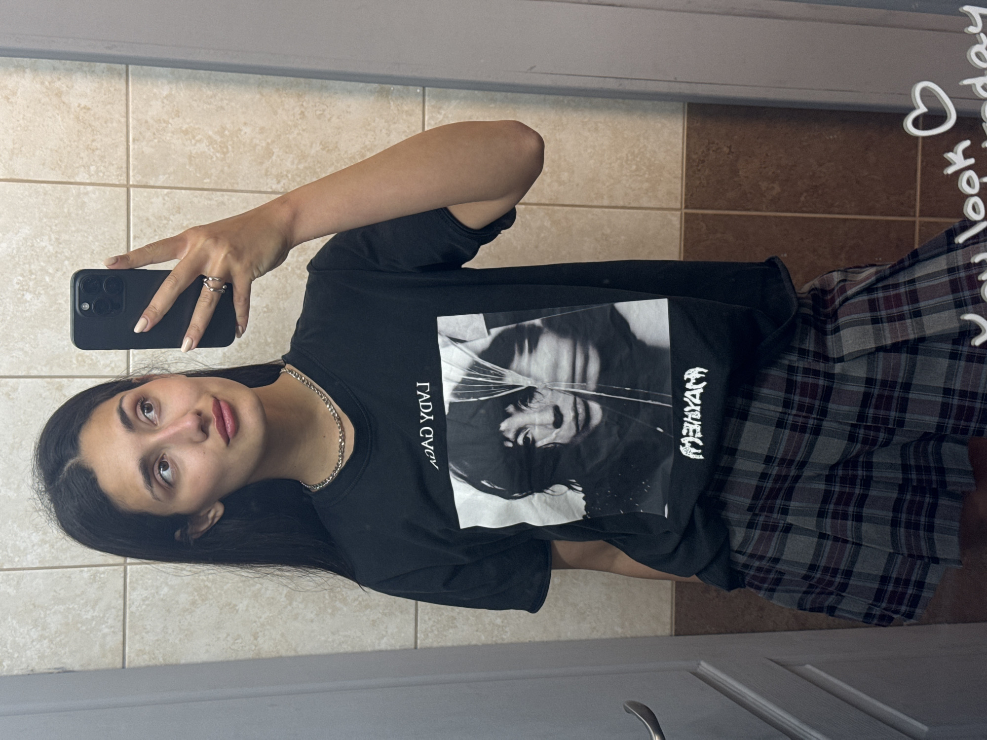 A young woman stands in a bathroom, holding a smartphone in her right hand and taking a selfie. She is wearing a black t-shirt with a black and white graphic print, a silver chain necklace, and a gray plaid skirt. Her long, dark hair falls over her left shoulder. The background is a bathroom with beige tile walls and floor. The lower portion of a white door is visible at the bottom. A message is written on the mirror, but the text is obscured. The location is likely not in Bar, Montenegro, based on the typical bathroom characteristics observed.