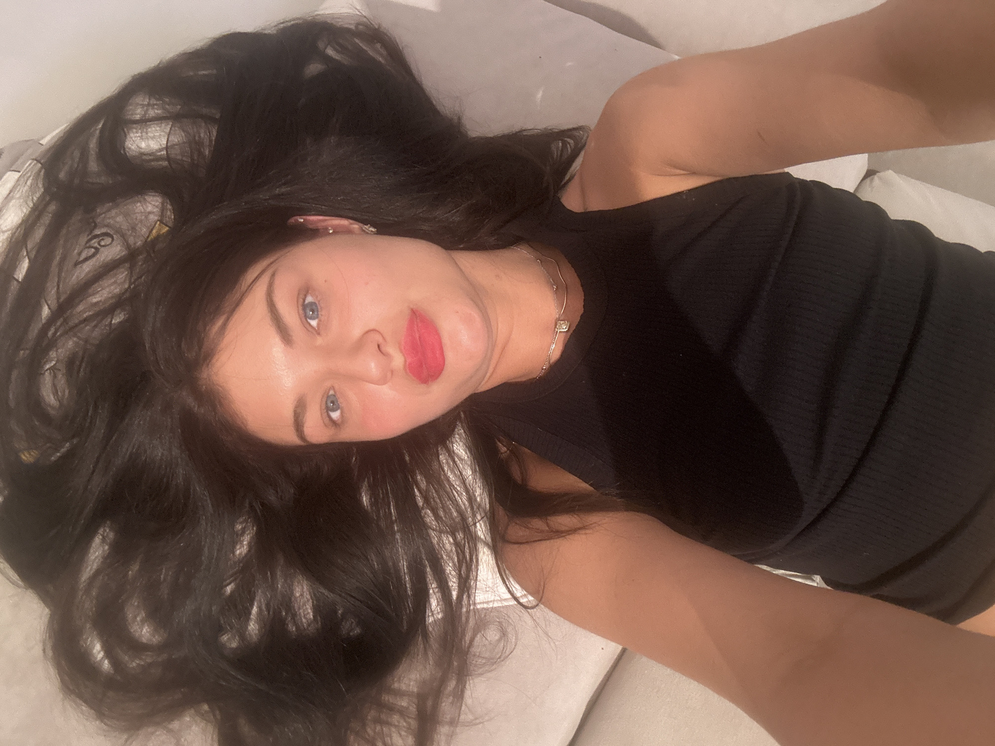 A young woman is captured in a selfie, lying down and looking directly at the camera. Her head is tilted to the left, with her long, dark, wavy hair fanned out around her against a light-colored, soft surface, likely a bed or sofa. She has piercing blue eyes, defined eyebrows, and is wearing vibrant red lipstick, with a neutral expression on her face.
She is dressed in a black ribbed tank top, and a delicate silver necklace with a small, light-colored pendant adorns her neck. Multiple ear piercings are visible on her left ear, including studs and a helix hoop. Her bare arms are visible, framing the lower part of the image. The indoor lighting is soft and even, creating a relaxed and intimate atmosphere. No visible text is present in the image. This scene is set in Moscow, Russia.