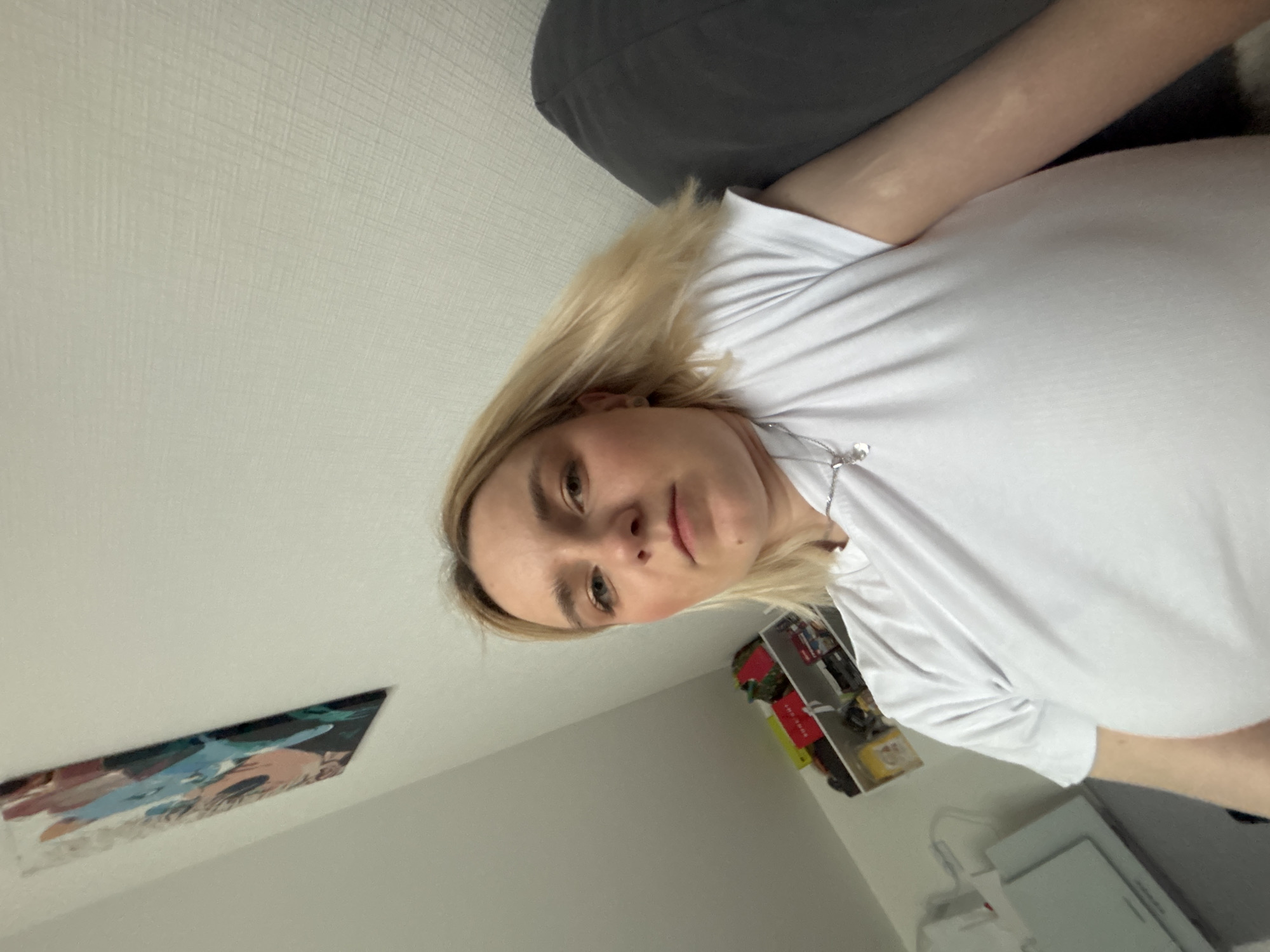 A young woman with blonde hair is lying on a gray pillow. She is wearing a white shirt and a silver necklace. Behind her is a white wall with a poster of an anime character, shelves with toys, and a white object resembling a printer. There is insufficient information to pinpoint the exact location within Vsevolozhsk, Russia.