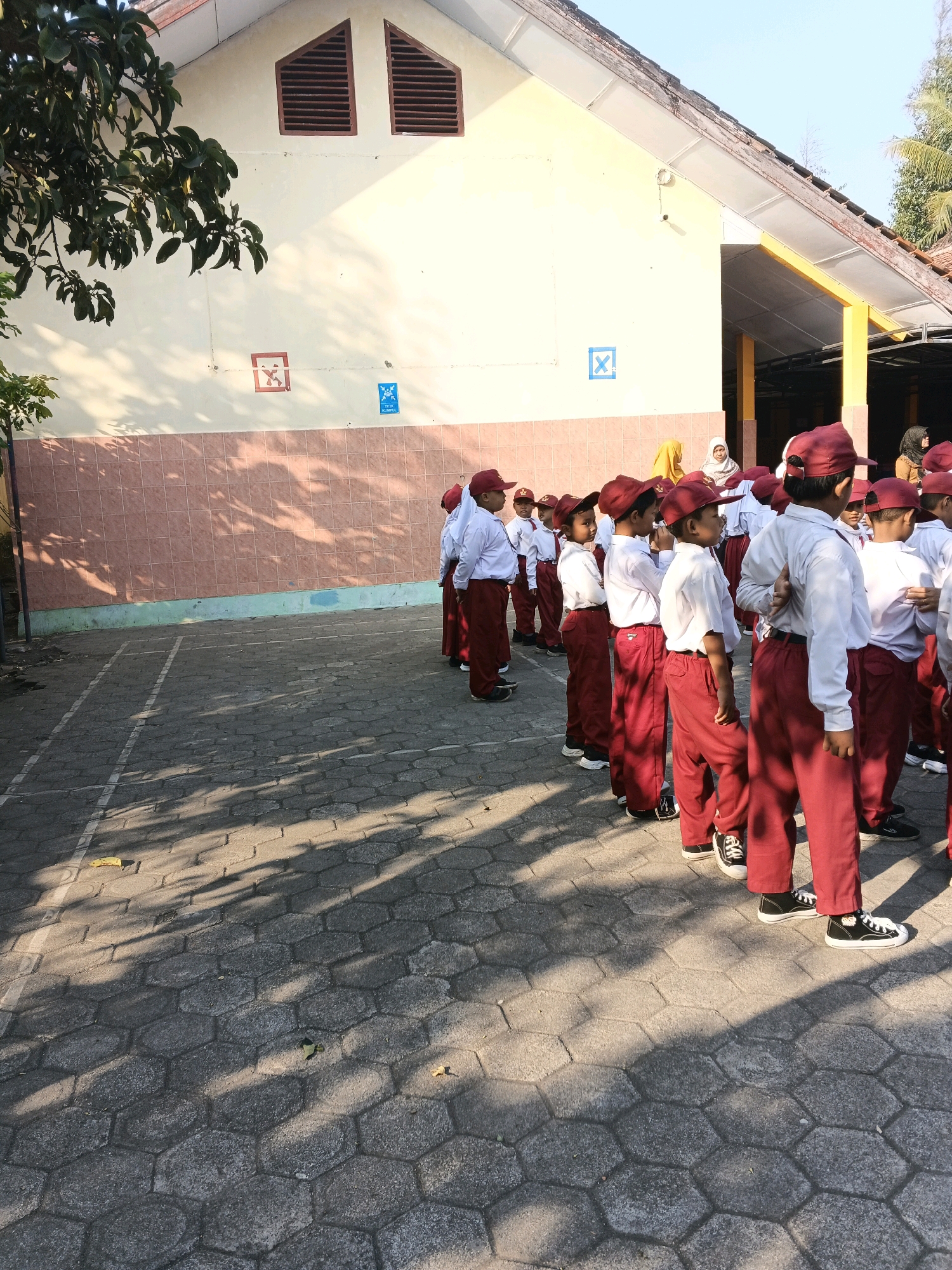 The image depicts a group of schoolchildren, likely in uniform, standing in a line on a paved area outside a building. The children are wearing red caps and maroon trousers, with white shirts. The building appears to be a school, with a yellow facade and a tiled roof. There are some markings on the wall of the building, including a red square with an 'X' inside and a blue square with a symbol that resembles a letter 'X' within a circle. The ground is paved with hexagonal interlocking stones, and the scene is illuminated by sunlight, casting shadows from trees and possibly the children themselves. The overall impression is that of an outdoor school activity or assembly. The location is identified as Glagah, Indonesia.