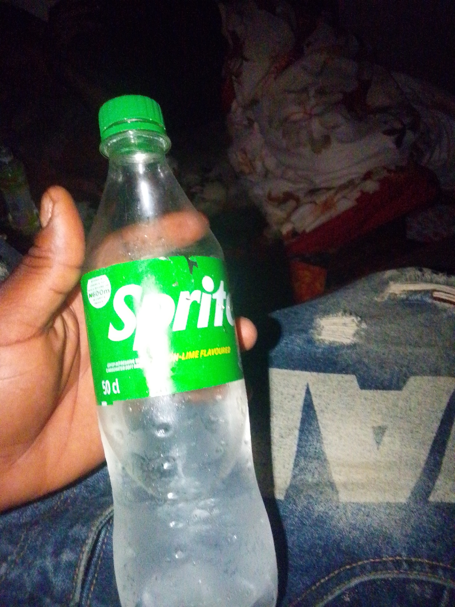 This image depicts a clear plastic 50 cl Sprite bottle, chilled and covered with condensation, held by a human hand with brown skin. The bottle features a green label with the "Sprite" logo in white text, indicating "LIME FLAVOURED," and "50 cl" volume. A green screw cap seals the bottle. A circular white emblem on the label displays "N600m." The individual holding the bottle is wearing blue denim jeans with visible distress marks and a light-colored, patterned design, partially showing geometric shapes resembling "WA" or similar elements. The background is dimly lit and out of focus, revealing patterned fabric, possibly a blanket or bedspread with floral or abstract designs in shades of white, brown, and red, and a solid reddish fabric beneath it. The scene is set indoors in the city of Keffi, Nigeria, under low light conditions.