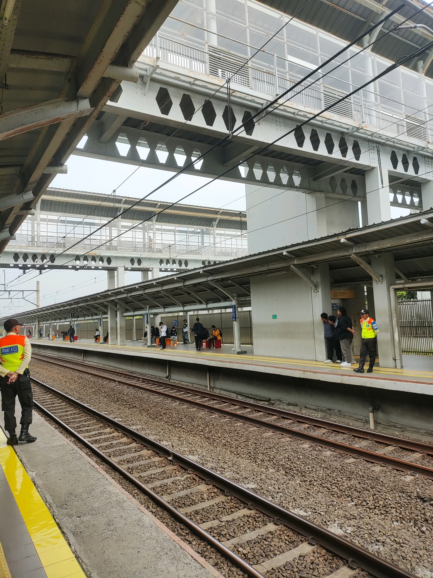 An outdoor view of a train station in Telagamurni, Indonesia, shows two sets of train tracks in the foreground. A platform runs along the right side of the tracks, with several passengers waiting. A man in a high-visibility vest and black pants, identified by the "PAM KCI" text on his vest, stands facing the camera on the platform. Further down the platform, two other individuals in similar vests are seen interacting with passengers. Above the platform, a covered walkway with transparent panels and structural beams is visible, connecting to an elevated pedestrian bridge. The sky is overcast.