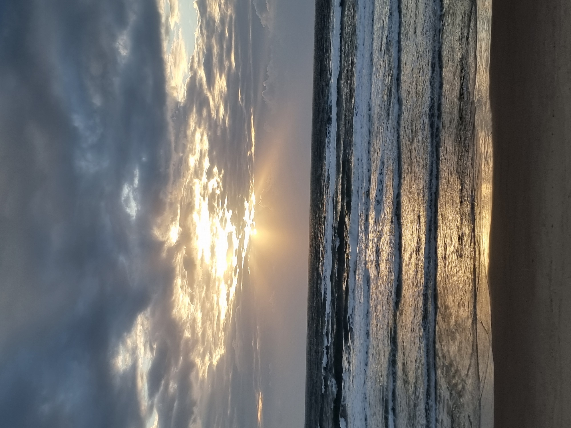 The image depicts a seascape at sunset. The sky, occupying the left half of the image, features dark, layered clouds with the sun shining through them. The ocean, on the right half, is characterized by distinct lines of breaking waves parallel to the shore. The beach is visible in the lower right corner. The sun's reflection is visible on the water. Based on the characteristics of the beach and the general geography visible, it is possible that the image was taken at a beach in Herzliya, Israel.