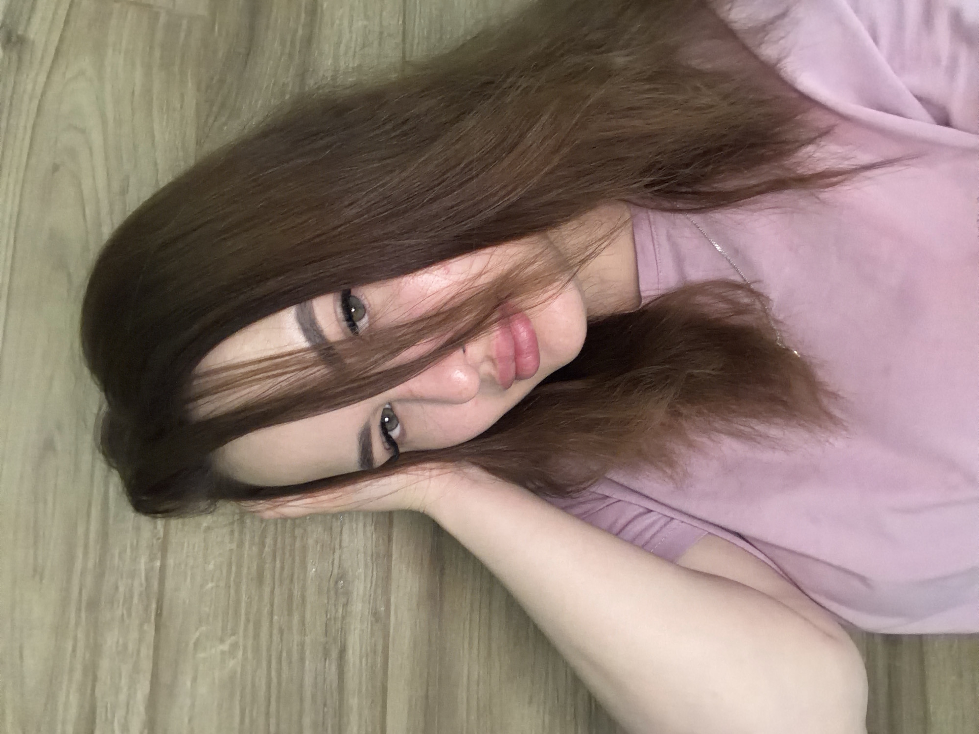 A young woman with brown hair and light skin is lying on a wooden floor. She wears a light pink t-shirt and a silver chain. Her head rests on her arm, with some of her hair partially covering her face. The floorboards run horizontally in the background. The location is likely indoors but cannot be identified as Krasnodar, Russia, without further information.