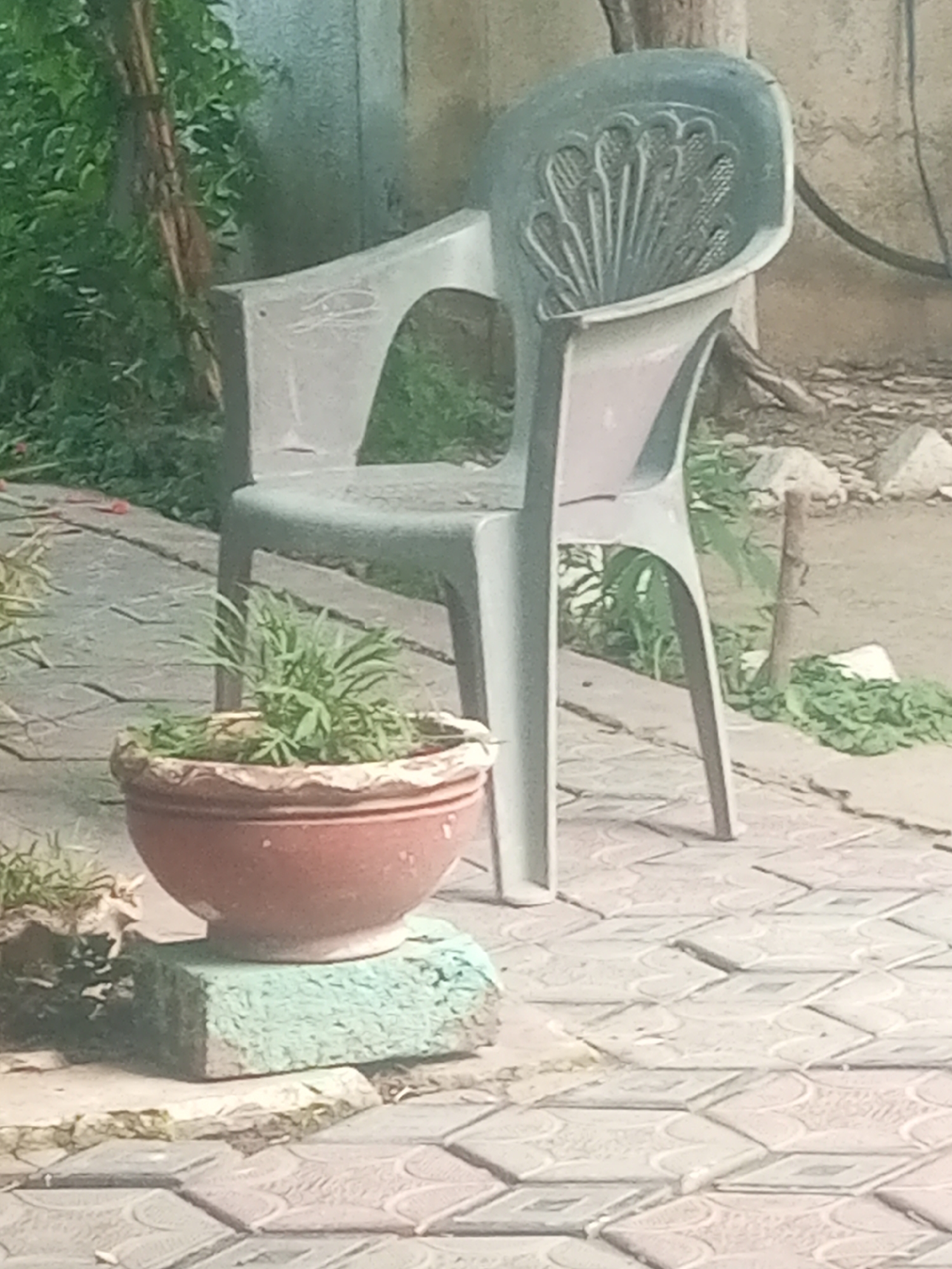 The image depicts an outdoor scene at a residence in Mansehra, Pakistan. In the foreground, a terracotta pot filled with green plants sits on a light blue, rectangular concrete block. This block is positioned on a paved surface composed of interlocking concrete tiles with a diamond pattern. Behind the pot and block stands a grey plastic chair with a decorative, fan-shaped design on its backrest. The chair is empty. To the left of the chair, dense green foliage and a vertical wooden pole are visible. The background shows a textured wall and more greenery. The lighting suggests it is daytime, with shadows present.