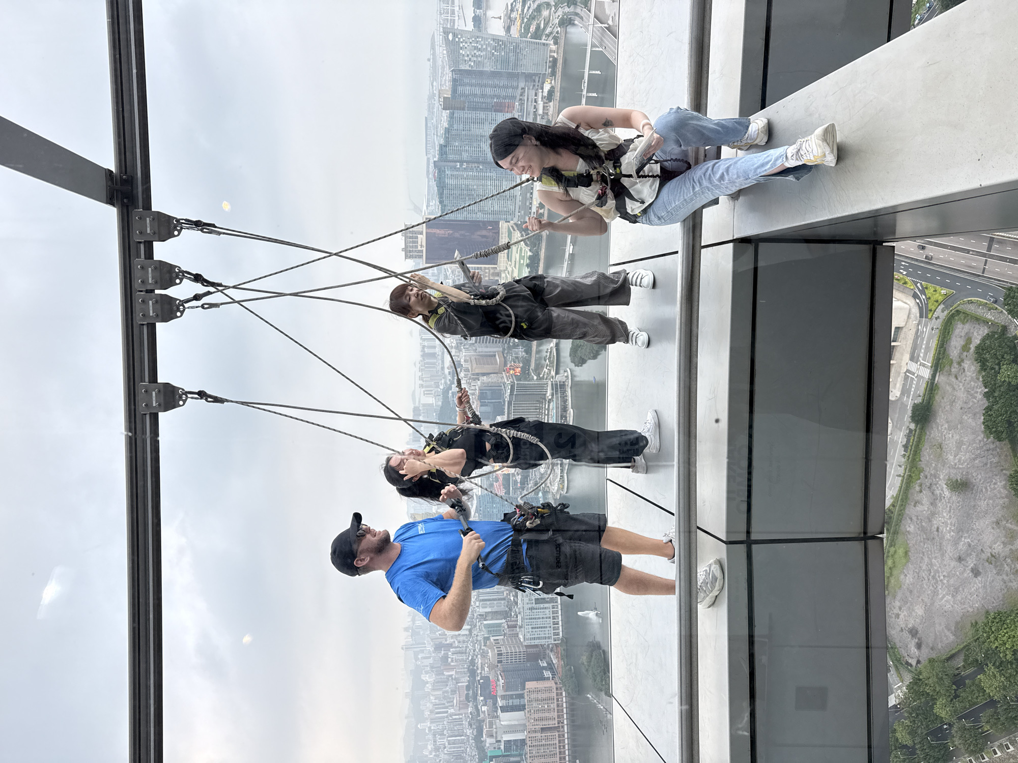 The image shows four people rappelling or doing a similar activity on the side of a tall building, with a cityscape visible in the background. The location is in Macau, China, based on the architecture and cityscape.
From bottom to top of the frame:
A man is standing, secured to the rappelling system, wearing a blue t-shirt, shorts, and a baseball cap. He is looking upwards, possibly instructing or monitoring the other participants.
Above him, two individuals are suspended horizontally, attached to the same rappelling system. They appear to be wearing harnesses and protective gear.
The fourth individual, at the top of the frame, is sitting on the edge of the building, with her legs dangling. She's also secured by the safety equipment.
The rappelling system consists of a metal structure attached to the building's exterior, with multiple cables connected to the individuals' harnesses. Below, the ground shows a mix of buildings, water, and green spaces. A highway is also visible. The weather appears overcast.