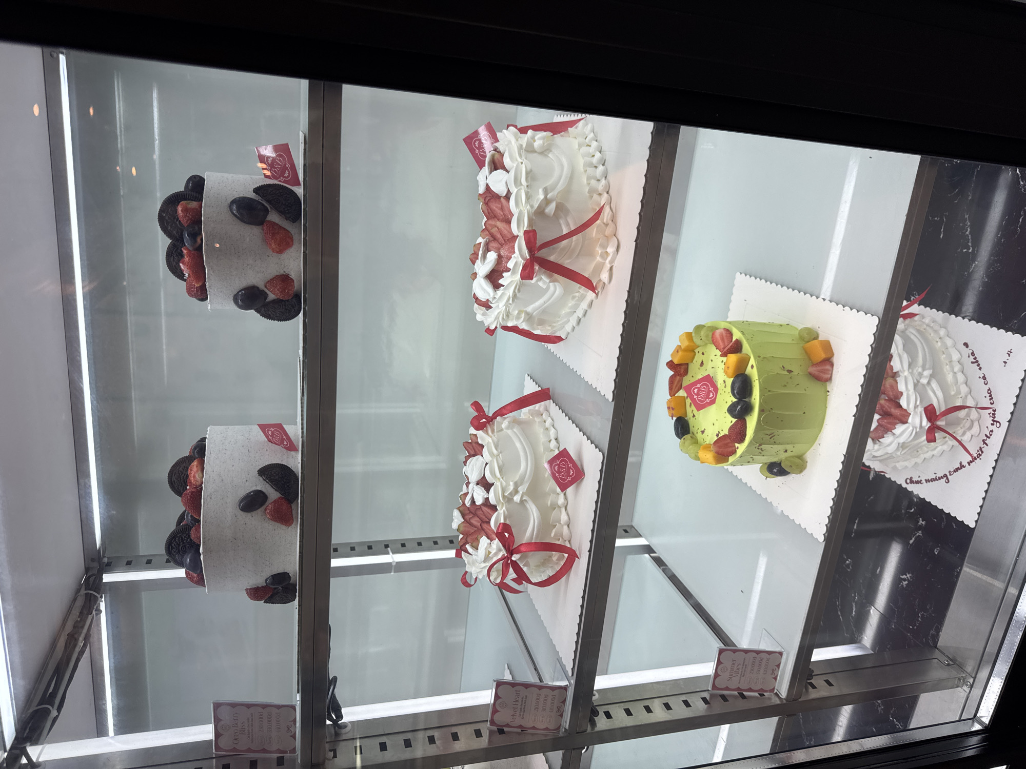 A commercial refrigerator displays four different frosted cakes, all on white doilies. The cakes are arranged on shelves, with two cakes on the top shelf and two on the second shelf. The first cake is white with dark cookies and red berries attached to the outside. The second cake is also white with a red ribbon around it and some red berries attached. The third cake is a light green color with yellow mangoes and red strawberries on top. The fourth cake is white, also with a red ribbon, and is set on a white doily that reads, "Chuc naing sinh nhật Ma yuu của cả nhà." Three different price tags appear under the cakes, stating the prices for each size. The price tags indicate that these cakes are in Đà Nẵng, Vietnam.