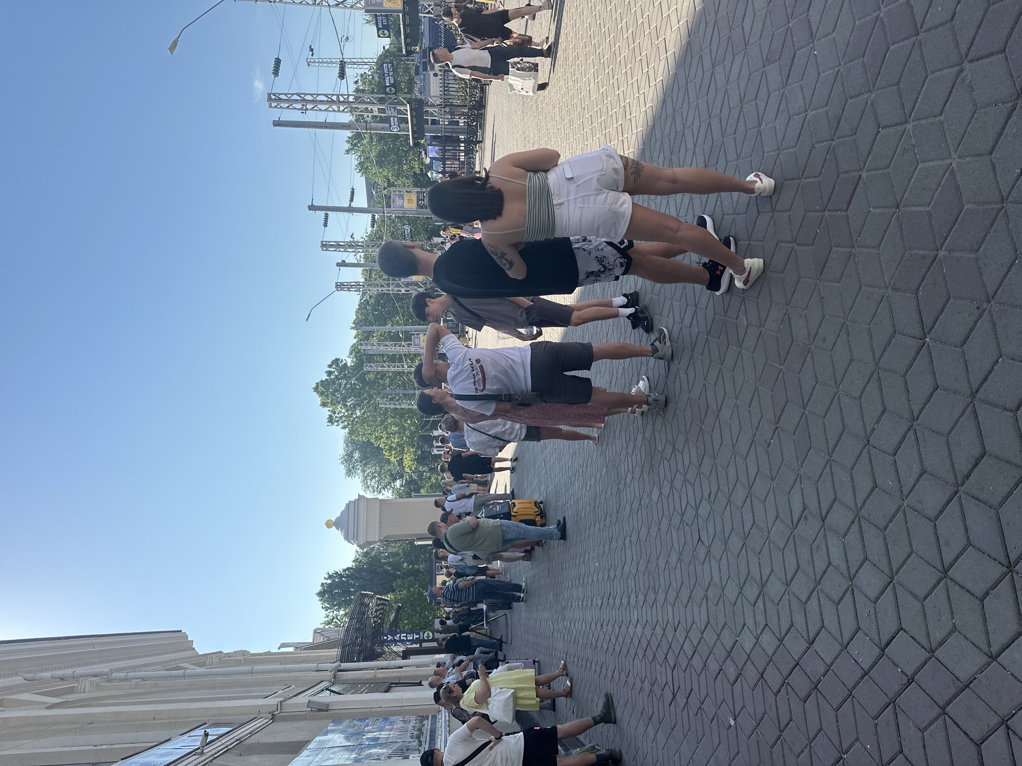 The image depicts a queue of people standing on a paved sidewalk outside a building with classical architecture. The building appears to be a public institution, possibly a train station or a cultural center, given the presence of signage in Ukrainian and English, including an "EXIT TO CITY" sign. The scene is set in Odesa, Ukraine, under a clear blue sky. People of various ages and genders are dressed in casual summer clothing, some are carrying luggage, suggesting travel. Electrical infrastructure, including poles and wires, are visible in the background, along with trees and a street lamp. The pavement is made of interlocking dark grey bricks. The overall impression is of a busy public space on a sunny day.
