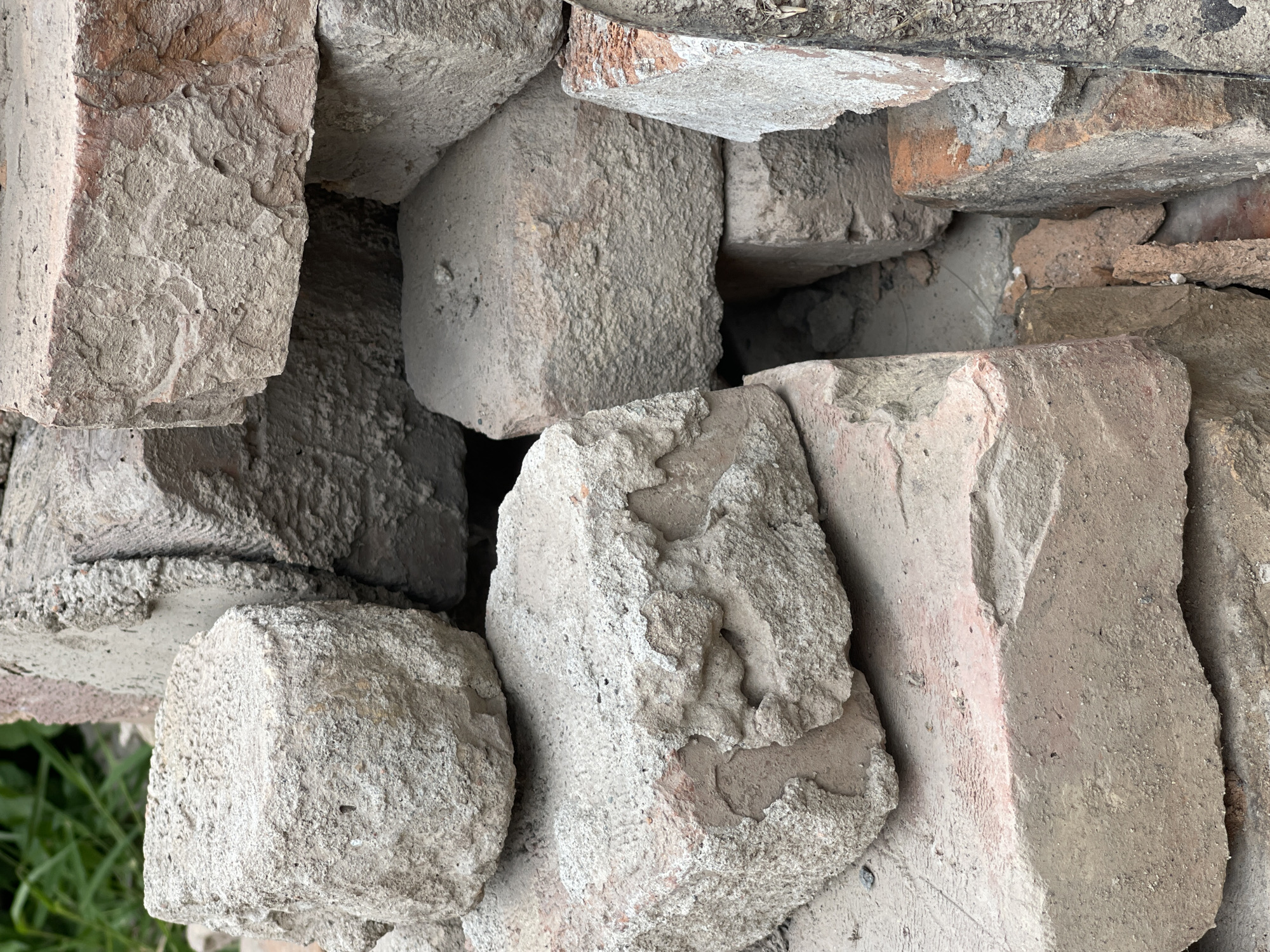 A close-up, eye-level, vertical shot showcases a pile of weathered, discolored, and broken bricks. The bricks exhibit varying shades of gray and brown, with some showing signs of red undertones, suggesting they may be made of clay or concrete. Their surfaces are rough, textured, and uneven, marked by chips, cracks, and the remnants of mortar. In the upper left quadrant, out-of-focus green foliage is visible, hinting at an outdoor setting. The lighting appears natural, casting subtle shadows that emphasize the three-dimensionality and aged condition of the bricks. The overall impression is one of disuse and decay. This scene could be located in an outdoor construction site, a demolition area, or a storage yard within Bishkek City, Kyrgyzstan.