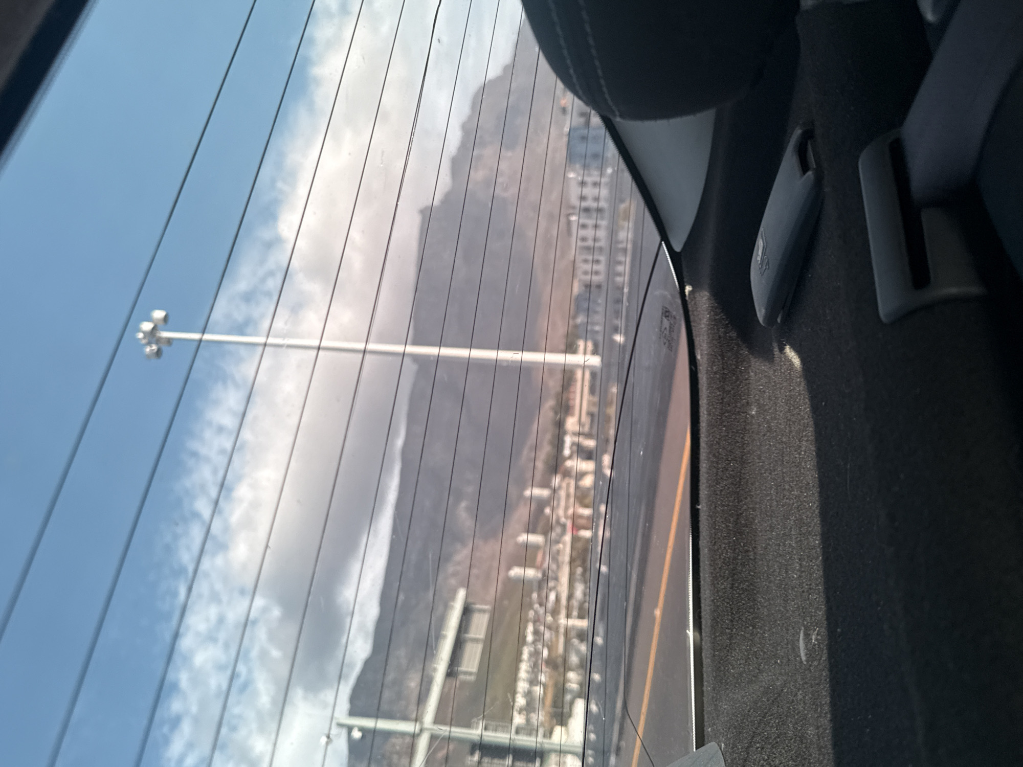 The image, taken from the interior of a vehicle through the rear window, shows a view of Cape Town, South Africa. The scene outside is partially obscured by the window's defroster lines. In the distance, Table Mountain is visible. Below the mountain, a cityscape with buildings and infrastructure stretches out. In the foreground, portions of the vehicle's interior, including a headrest and seatbelt mechanism, are visible. The sky is blue with scattered clouds.