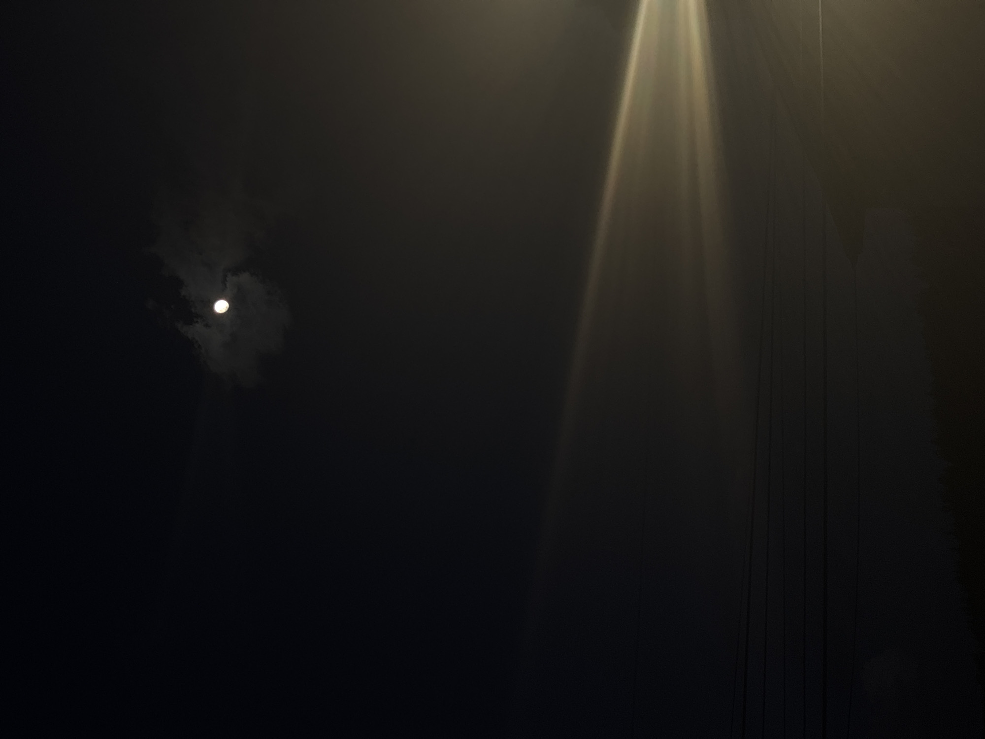 A nighttime scene depicts a faint moon partially obscured by clouds in the upper left. To the right, streaks of light emanate downwards, possibly from a light source outside the frame. Vertical lines are visible on the right, possibly wires or cables. The scene is predominantly dark, obscuring any further details about location or objects. No people are visible. There is no evidence in the image to confirm the location is Borino, Bulgaria.