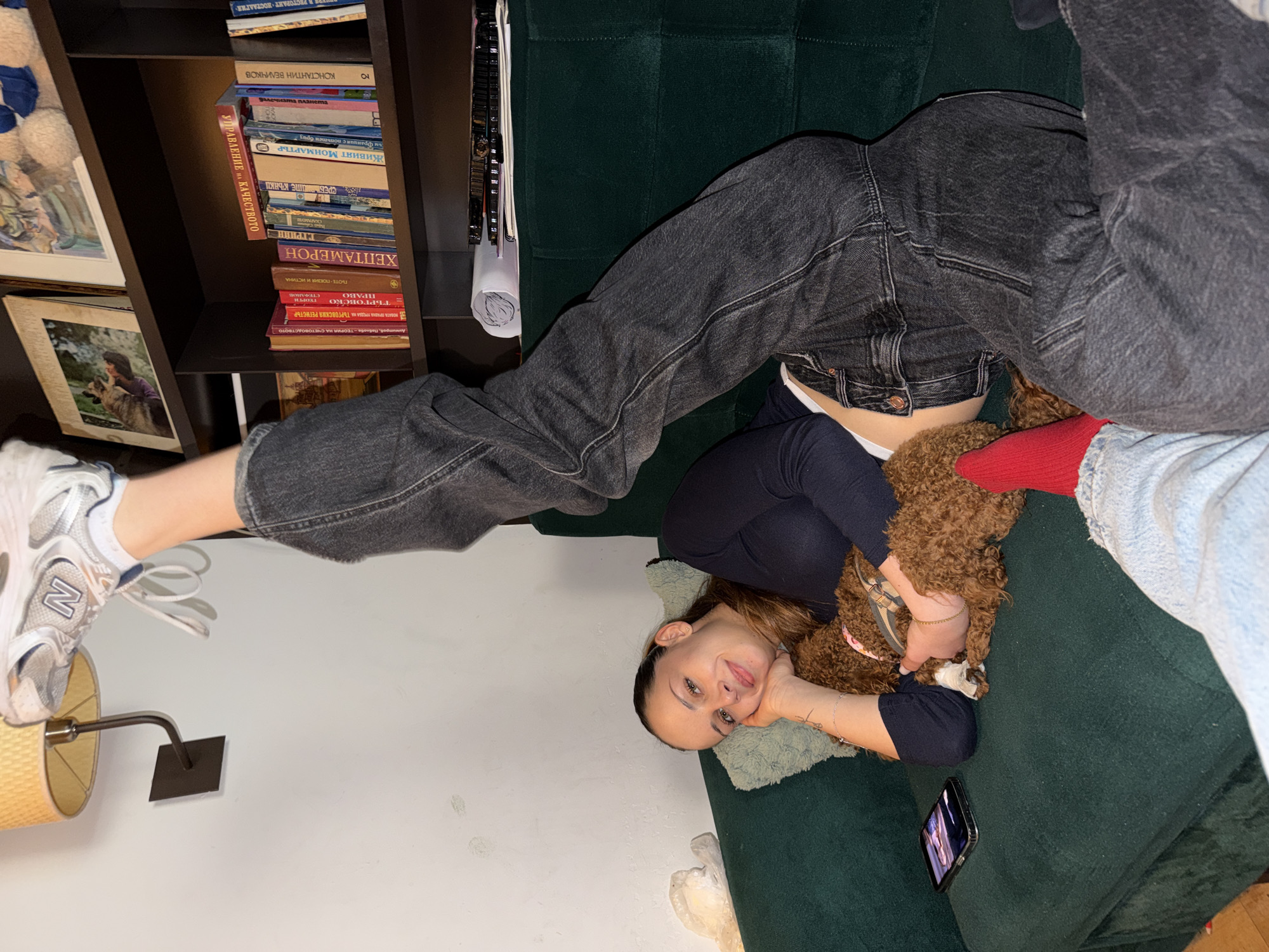 A young woman in a dark top and dark jeans is lying upside down on a green sofa, with one leg straight up in the air wearing a sneaker. A brown dog is lying on her stomach. A phone is on the sofa next to her. Bookshelves with books in Bulgarian are in the background. A lamp with a beige shade is to the left. The location is likely Sofia, Bulgaria, given the books in Bulgarian.