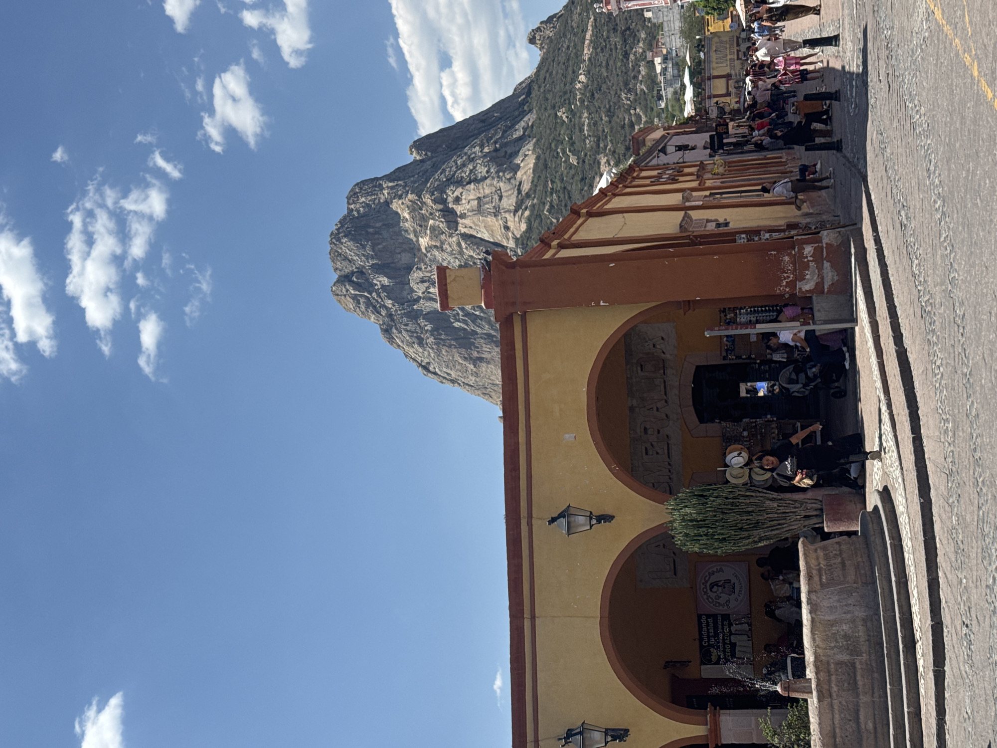 The image shows a street scene in Bernal, Mexico, with a view of Peña de Bernal (Bernal Peak) in the background. The sky is blue with scattered white clouds. In the foreground is a section of a building with arched openings, painted in shades of yellow and orange. People are walking on the cobblestone street next to the building. There's a fountain and some vegetation along the building's base. Some of the arched openings appear to house shops or vendors. The cobblestone street is lined with buildings and structures in the distance.