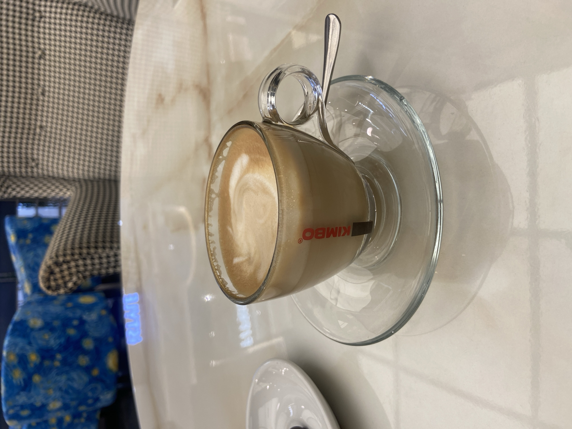 A clear glass cup filled with coffee and foam sits on a glass saucer on a polished marble table. A spoon rests on the saucer, partially beneath the cup. To the left of the coffee cup is a small white saucer with a dark object, possibly a piece of chocolate or a berry. In the background, a chair with a black and white houndstooth pattern is visible, and further back, a blue chair with a starry pattern is partially in frame. The lighting suggests an indoor setting, possibly a cafe or lounge area. The scene is devoid of people.