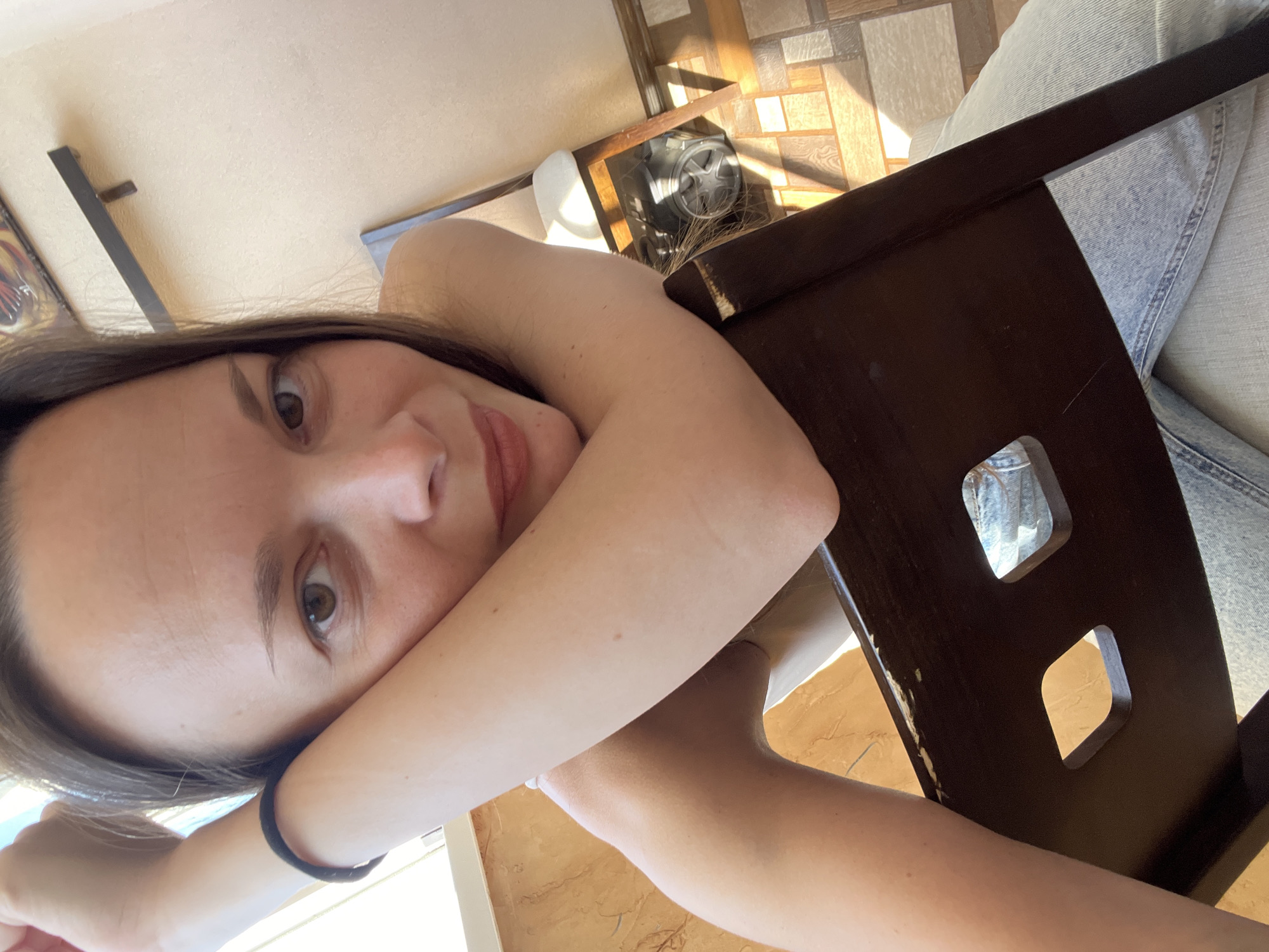 A woman with long, dark hair is pictured in a selfie, resting her arms on the back of a dark brown wooden chair. She is wearing a light-colored top and blue jeans. Her left arm is bent, with her hand resting near her face. A black hair tie is visible on her left wrist. The background includes a tiled floor, a wall with a shadow cast by sunlight, and a framed picture on the wall. Underneath the chair, a portion of a vacuum cleaner is visible. The scene appears to be indoors, possibly in a home or apartment. There is no specific indication of its location within the city of Tolyatti, Russia.