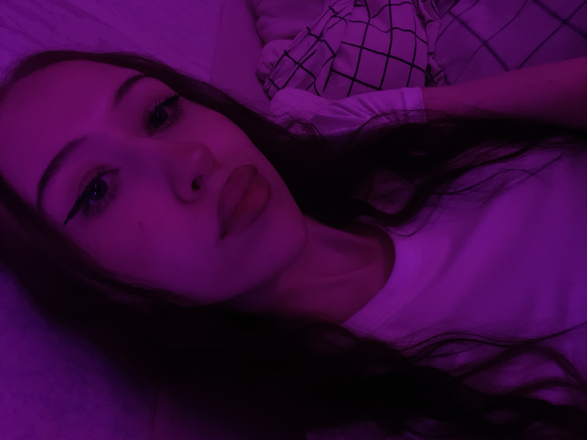 This is a close-up, first-person perspective selfie of a young woman lying down. The scene is dimly lit with a strong purple hue, suggesting artificial lighting or a nighttime setting. The woman is looking directly at the camera with her eyes open and a subtle makeup application, including eyeliner. Her lips are parted, with a noticeable pout. Her dark hair is spread out around her face and shoulders. She is wearing a white t-shirt. In the background, a person's arm is visible, and a crumpled blanket with a black grid pattern can be seen. The overall mood appears relaxed and intimate, possibly taken in a bedroom or a comfortable indoor space. There is no visible text.
