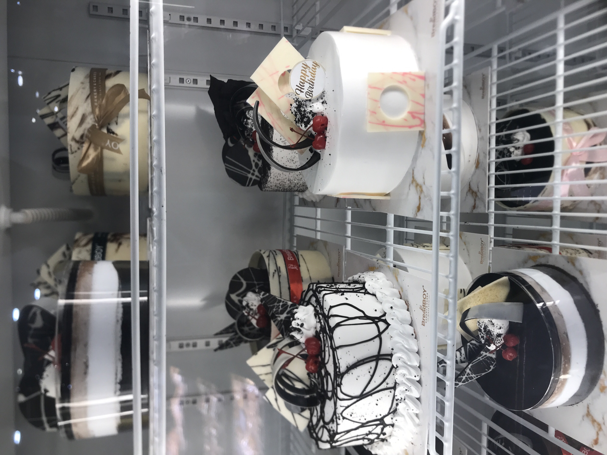 The image shows a refrigeration display case filled with various cakes in a bakery. The cakes are presented on white shelves, and the case itself is made of metal and glass.
On the top shelf, there are two cakes. The cake on the left is partially obscured, but appears to be a layered cake in a clear plastic container with dark and white layers. The cake on the right is round with a marble-like frosting and decorated with a gold ribbon and chocolate shavings. A label on this cake reads "BAKERY & CAKES".
The middle shelf features two more cakes. The cake on the left is round and white with black icing drizzled across the top, adorned with cherries, white frosting swirls, and chocolate decorations. The cake on the right is also round and white, with a "Happy Birthday" topper, chocolate and cream decorations, and square white chocolate pieces with pink accents.
The bottom shelf contains two more cakes. The cake on the left is a dark chocolate cake with white and brown layers visible through a clear plastic wrapping, decorated with cherries and chocolate elements. The cake on the right is partially visible, with a pink bow tied around its middle.
The environment appears to be a bakery, and the display case is illuminated by overhead lights. The overall impression is of a well-stocked bakery ready for sale.
