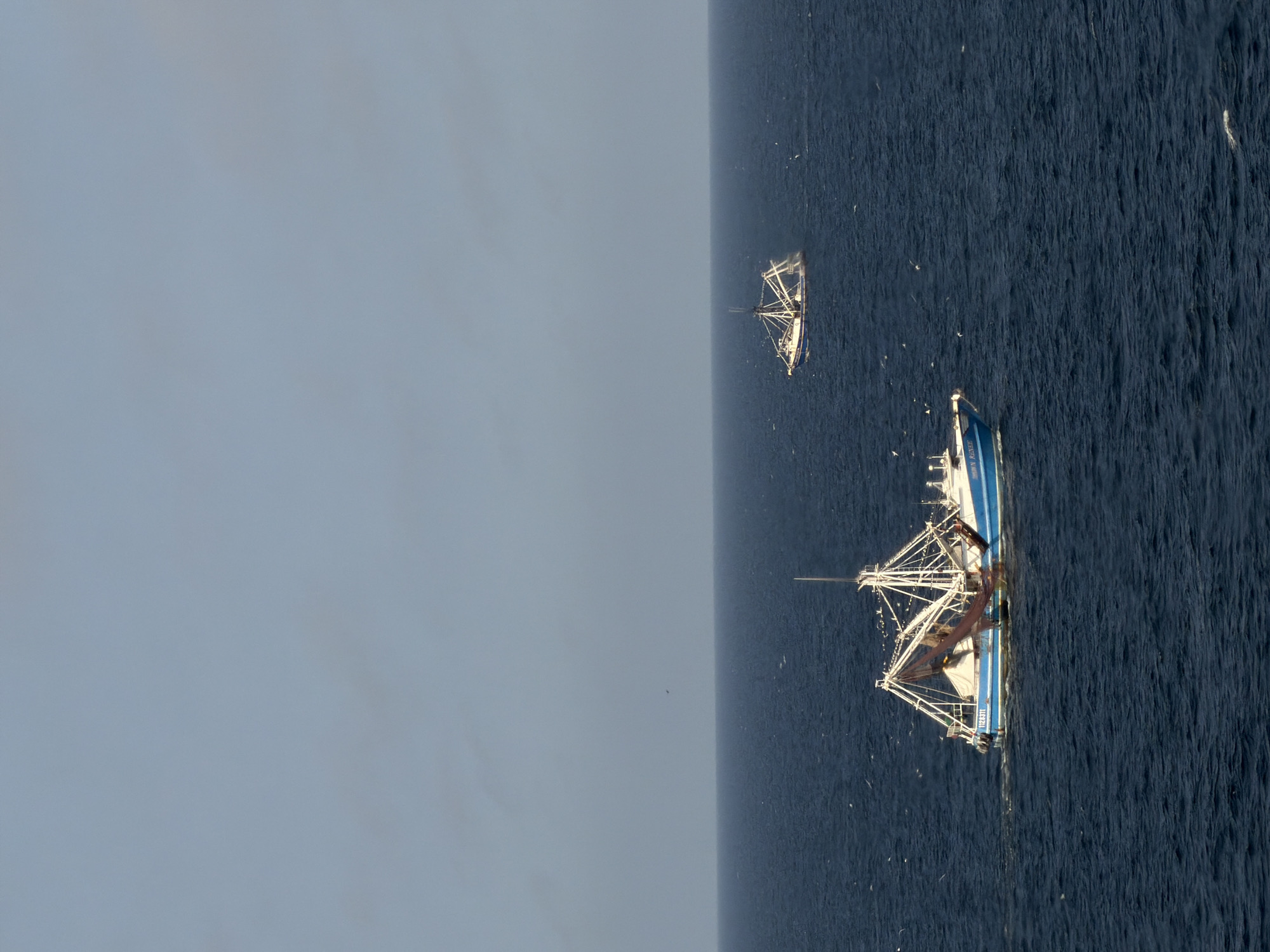 The image, captured from an aerial perspective, shows two fishing trawlers operating on the open ocean under an overcast sky. The photograph is oriented 90 degrees clockwise, placing the horizon vertically in the frame, separating the deep blue, rippled water on the right from the uniform light gray sky on the left.
In the foreground, a larger fishing boat, predominantly white with a prominent blue stripe along its hull, is visible in the lower right portion of the image. Its fishing gear, including large outriggers and what appear to be trawl doors and nets, is deployed, extending out from both sides of the vessel. A wake is visible behind the boat, suggesting it is in motion. On the stern, the number "1122471" is clearly discernible, with partially obscured text below it. The bow also features a blue stripe with illegible white text.
Further in the distance, towards the upper left, a second, smaller fishing trawler is seen. It also has its fishing equipment, including masts and booms, extended, indicating active fishing. Both boats appear to be engaged in trawling.
The water surface is covered in small ripples, and several small white specks, possibly seagulls or sea spray, are scattered across the water around the vessels. The lighting is soft and diffused, characteristic of an overcast day, with no direct sunlight or shadows visible. No land is in sight, emphasizing the expansive nature of the ocean.