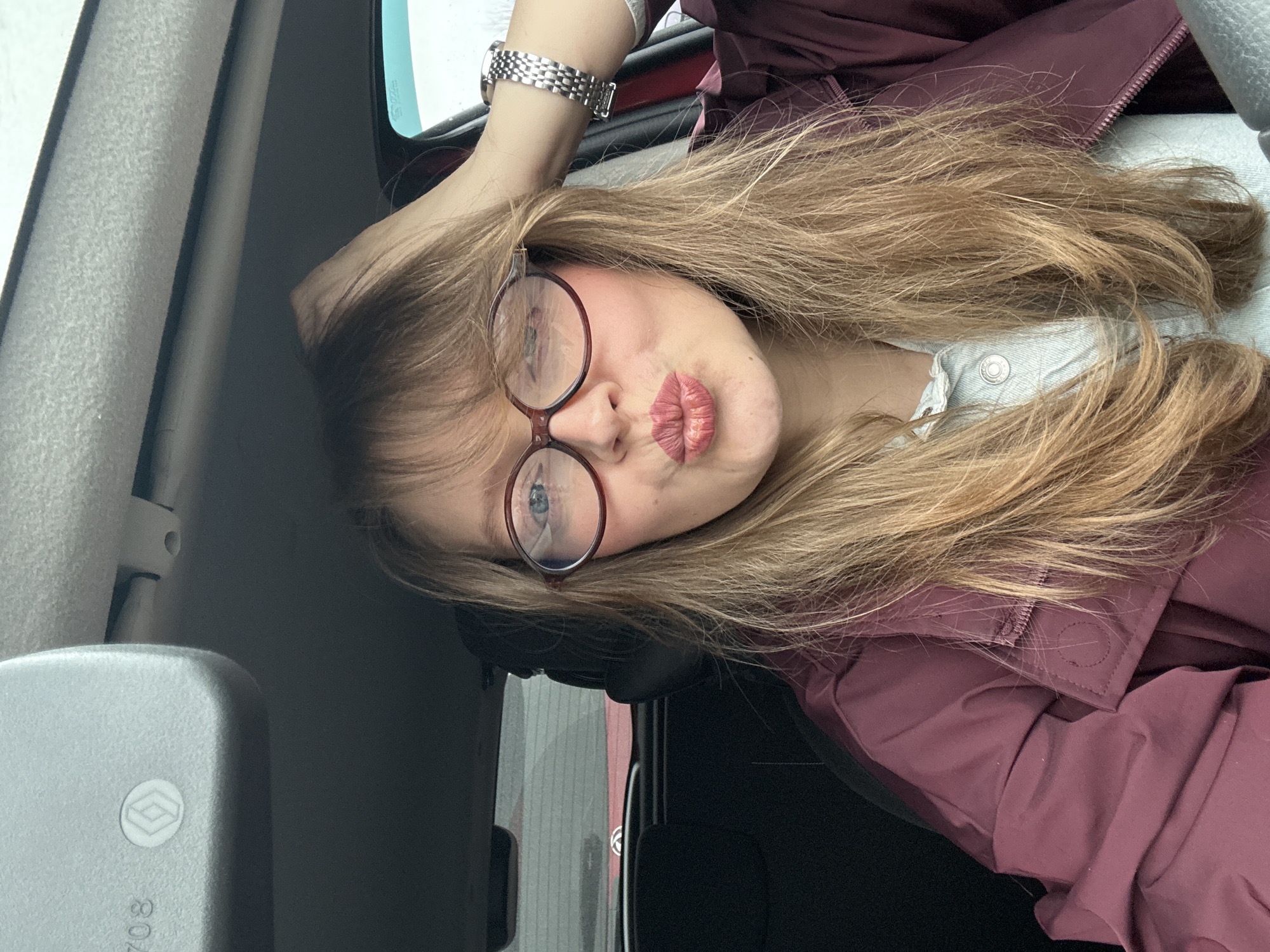 A young woman with long blonde hair and glasses is posing for a selfie in what appears to be the interior of a vehicle. She is wearing a maroon jacket over a light-colored collared shirt. The woman is wearing a silver watch on her left wrist, which is resting on the vehicle's interior. The background shows the interior of the vehicle and a glimpse of what might be the exterior environment through the window. There are no explicit cues pointing towards the city of Izhevsk, Russia.