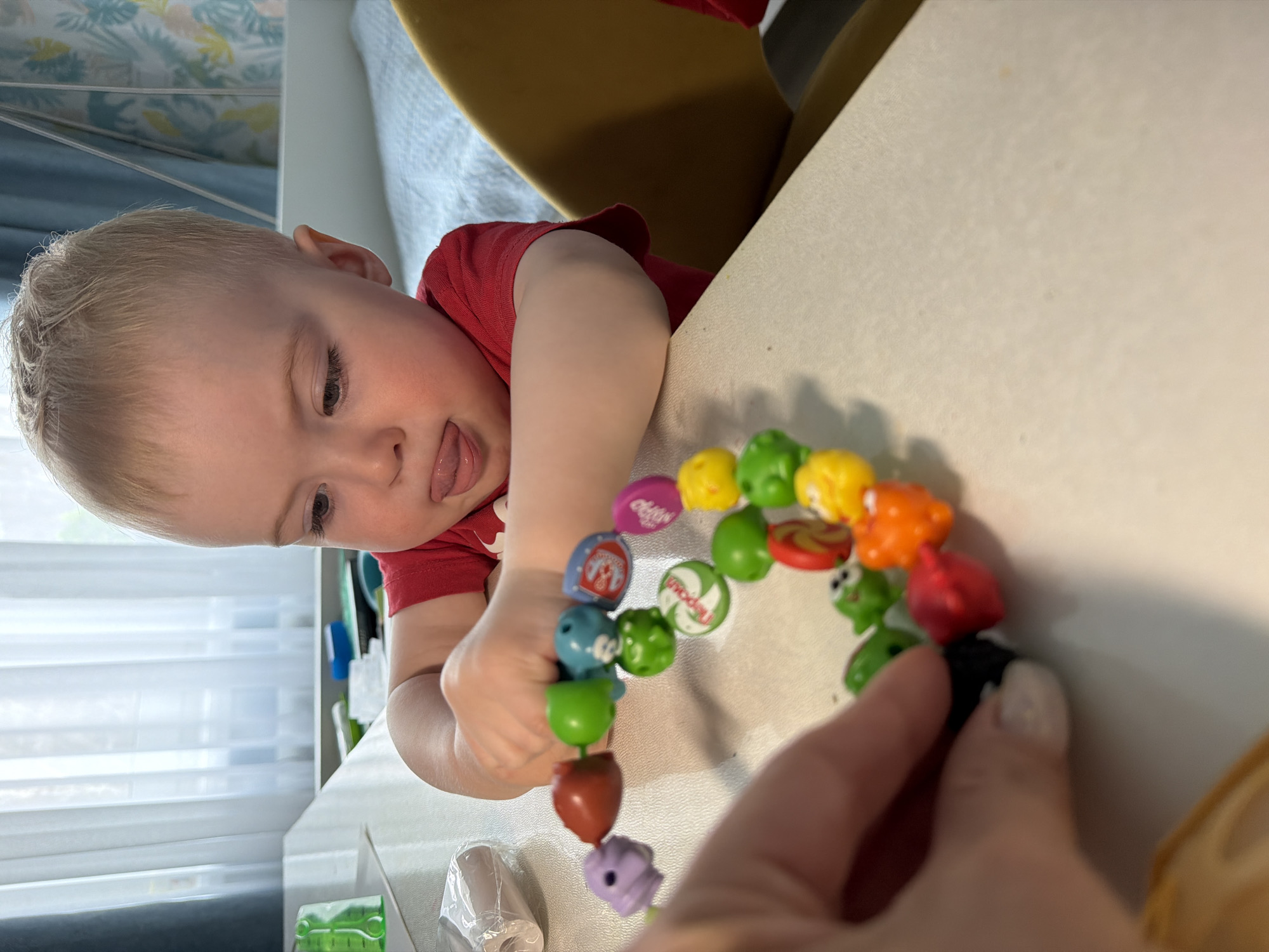 A young child with blond hair, wearing a red shirt, is positioned at a table. The child has his tongue slightly sticking out. He is holding a colorful string of beads. A hand is also holding the string of beads. The table is light-colored. The background includes a window with blinds, a chair with a yellow back, and a glimpse of a bed. It is impossible to verify the location of Votkinsk, Russia in the image.