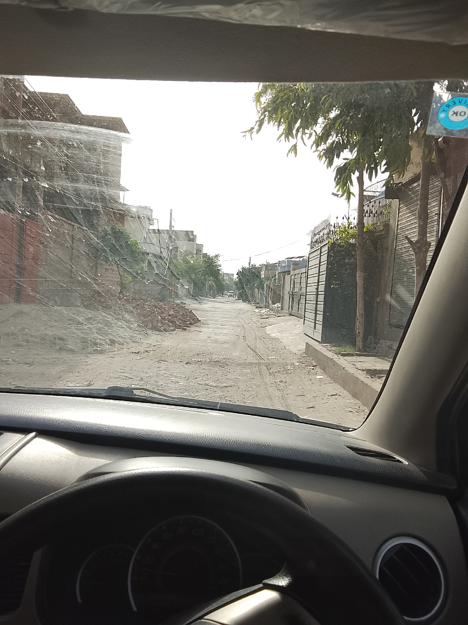 The photograph is taken from inside a car, viewing a dirt road in Gulgasht Colony, Pakistan. The road is lined with buildings on either side, some under construction or repair. A pile of bricks is visible on the left side of the road. The view through the windshield is partially obstructed by what appears to be water or debris. A sticker with the word "OK" is visible on the right side of the windshield. The car's dashboard and steering wheel are prominent in the foreground.
