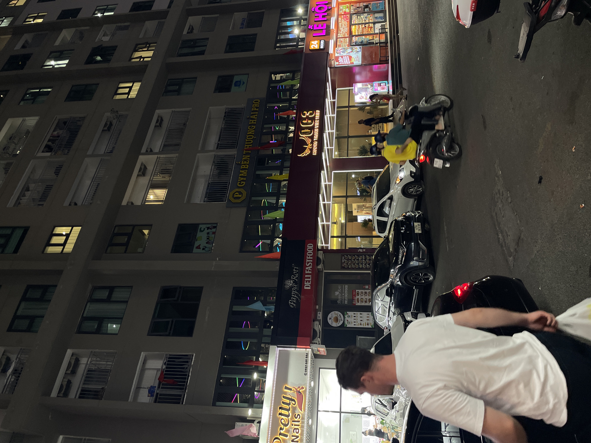 This image captures a street scene at night in Nha Trang, Vietnam, likely in a commercial area with a multi-story building housing various businesses on the ground floor. The building's facade features multiple windows and balconies, with some lights illuminated. On the ground level, a row of storefronts is visible, including "Pretty! Nails," "Pappa Roti DELI FASTFOOD," and "LÊ HỘI." A sign for "GYM BÊN THƯƠNG HẢI PRO" is also present above the businesses.
Several vehicles are present on the street, including cars and a scooter carrying two people. A man in a white t-shirt walks away from the camera, carrying a white plastic bag. Other individuals are seen walking or standing near the businesses. The lighting suggests it's evening or night, with the storefronts illuminated by neon and other artificial lights, creating a vibrant atmosphere. The street appears to be paved, and there are some debris on the ground.