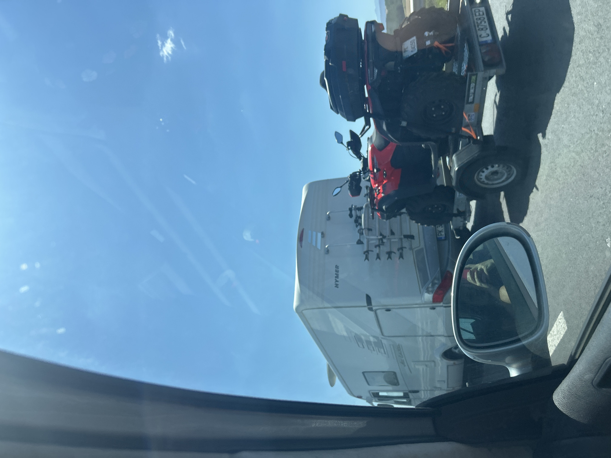 The image depicts a view from inside a vehicle, looking outwards at a sunny sky and a road. In the foreground, a silver side-view mirror reflects the face of the person taking the photo, who is holding a phone. To the right of the mirror, a trailer is visible, carrying two red ATVs. The ATVs are equipped with black storage boxes on the rear. Behind the trailer, a white recreational vehicle (RV) with the brand name "HYMER" is parked on the right side of the road. The RV appears to have a satellite dish mounted on its roof and a bicycle rack on the back. Further down the road, other vehicles are present. The environment suggests a rural or suburban setting, possibly a highway, given the presence of multiple vehicles and the open sky. The license plate on the trailer reads "C 8758 EP". The license plate on one of the ATVs is partially visible and appears to be "PK 04250". The scene is consistent with a road trip or travel scenario. The location is stated to be the city of Vakarel, Bulgaria.