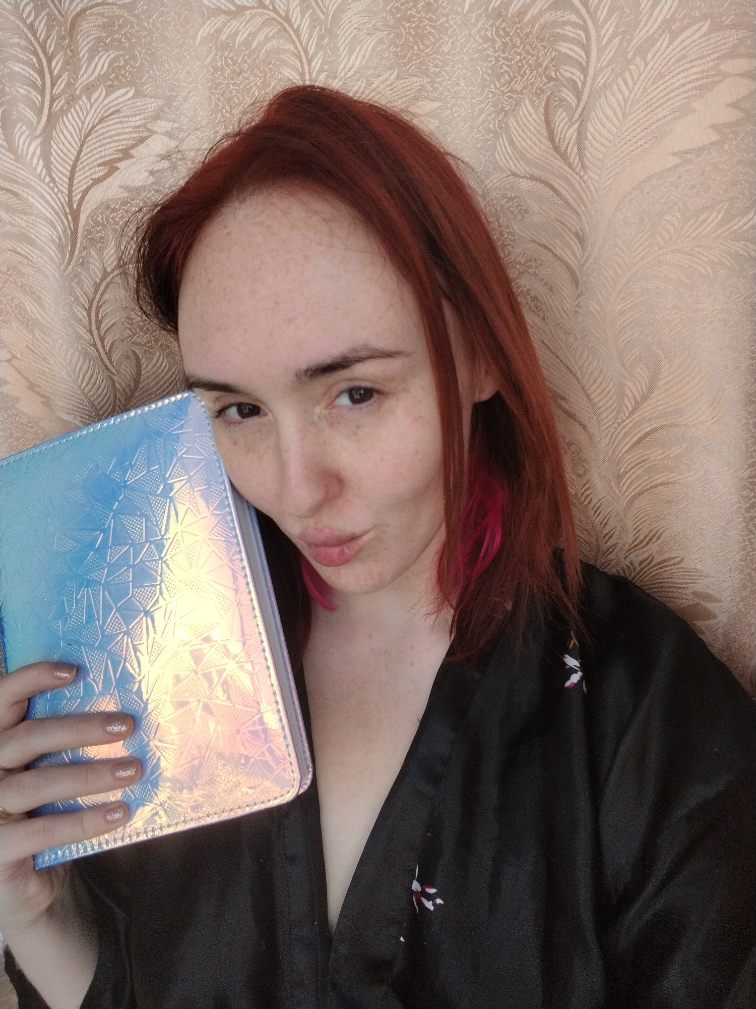 A woman with fair skin and freckles is holding a blue holographic-patterned book in her left hand. She has reddish-brown hair with pink streaks in it, and is wearing a black robe with a floral pattern. She is looking towards the camera and has slightly pursed lips. The background is a light beige patterned wall. There is no indication of Izhevsk, Russia in this image.