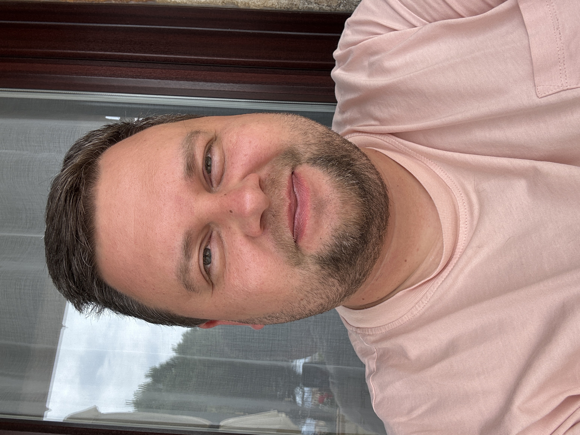 A fair-skinned man with short dark hair and a beard is looking at the camera with a slight smile. He is wearing a pink t-shirt. Behind him, a window is visible, reflecting an exterior scene. The location is likely Sant Feliu de Guíxols, Spain, based on previous information. Specific details about the exterior scene are not discernible.