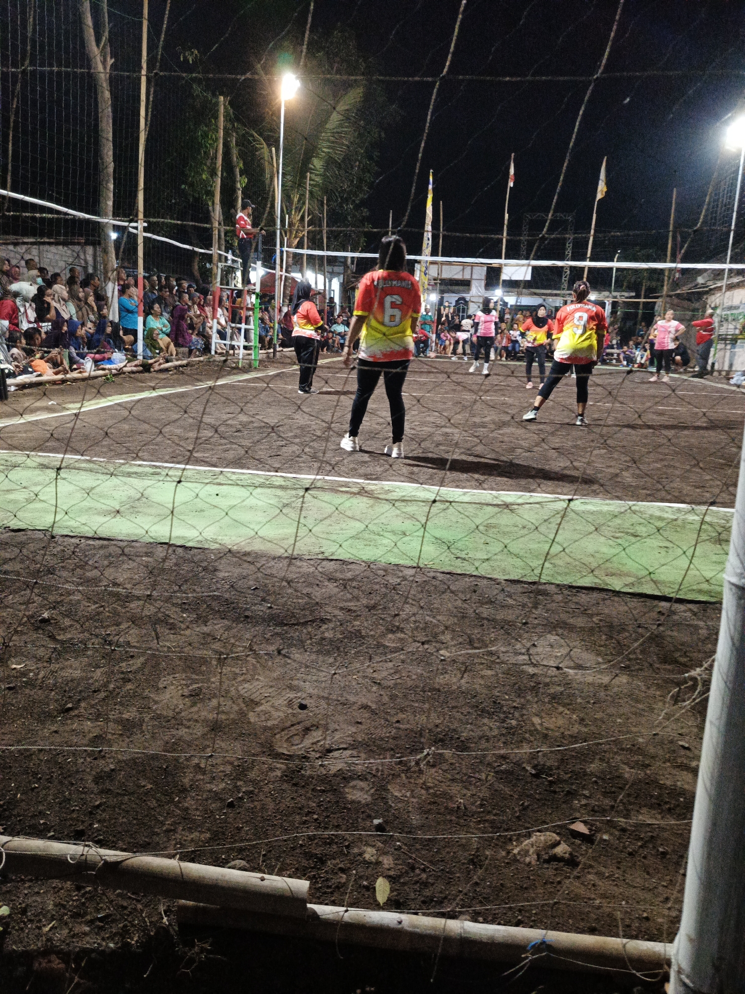 In the city of Rowokangkung, Indonesia, a volleyball game is underway at night, illuminated by artificial lights. The court, made of dirt and marked with white lines, is bordered by a net. Spectators are seated on the ground to the left of the court, many of them women dressed in casual attire. Several women, wearing athletic uniforms, are actively playing volleyball. One player, distinguished by the number 6 on her back and a yellow and red jersey, is the focal point. Another player, with the number 9 on her back and a similar uniform, is also visible. The scene is further enclosed by a fence and bamboo poles, with trees and vegetation in the background. The atmosphere appears lively, with the ongoing match and the presence of an audience.