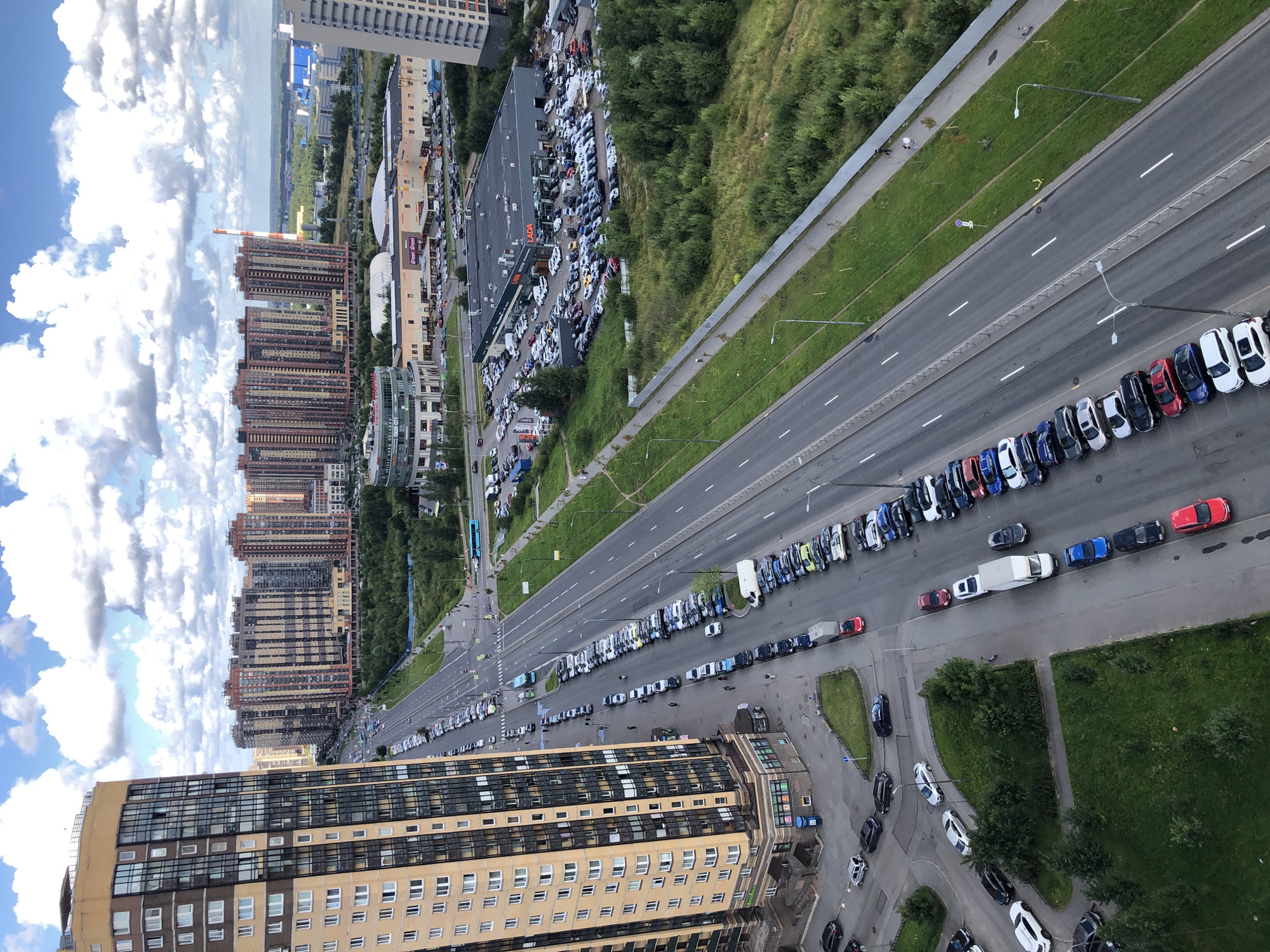An aerial, high-angle shot captures a sprawling urban landscape under a cloudy sky with patches of blue. In the foreground, a tall, multi-story residential building with a tan facade and dark windows dominates the left side of the frame. A wide, multi-lane road runs parallel to the building, filled with numerous parked cars on its right side. Further down the road, traffic is visible, including several buses and cars in motion.
Beyond the road, a dense green area of trees and grass separates the residential buildings from a commercial zone. This zone features several large, flat-roofed buildings, one of which displays the "LADA" logo, indicating a car dealership. A large parking lot in front of the LADA dealership is packed with cars. In the distance, more residential high-rises and industrial buildings can be seen, extending to the horizon under the vast sky. The overall scene depicts a modern, developing urban environment with a mix of residential and commercial infrastructure.
