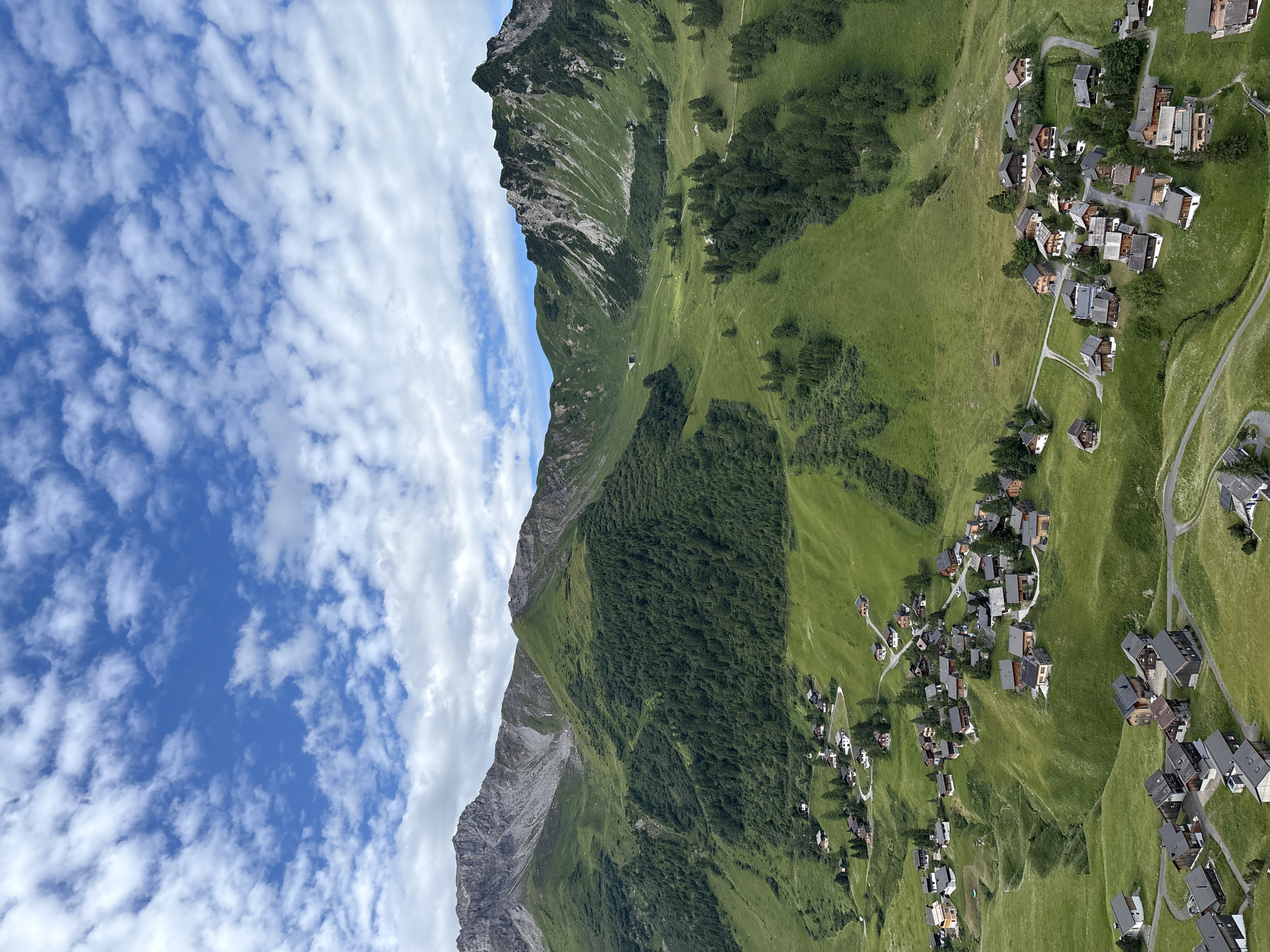 The image depicts a panoramic view of a mountainous landscape in Malbun, Liechtenstein. The scene is dominated by lush green hills and valleys, dotted with a cluster of small, traditional chalets. The chalets are predominantly wooden with dark roofs and many have balconies. Winding roads traverse the grassy slopes, connecting the scattered houses. In the background, imposing grey mountains rise, some with rocky peaks and others covered in dense green forest. The sky above is a vibrant blue, interspersed with scattered white clouds. The overall impression is one of a serene and picturesque alpine village.