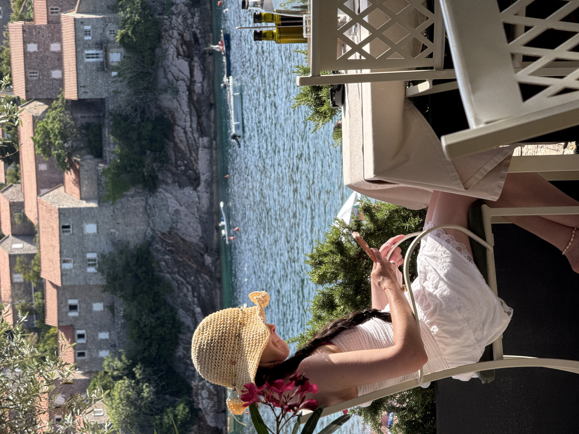 This image depicts an outdoor dining or relaxation scene at Sveti Stefan, Montenegro, with a woman seated in the foreground. The background features several multi-story stone buildings with red-tiled roofs, characteristic of the historic village, built into a rocky hillside overlooking a calm, blue-green body of water. Multiple small motorboats are visible on the water.
In the mid-ground, a woman is seated in a light-colored outdoor chair, viewed from her left side. She wears a wide-brimmed, light-colored straw hat and a white strapless dress with a textured bodice and a lace-trimmed hem. Her long, dark hair is styled in a braid or plait, and pink flowers are tucked near her left ear. She is holding a light-colored smartphone with both hands and looking down at its screen.
To the right of the woman, a square table is covered with a light-colored tablecloth. On the table are a dark green bottle (likely olive oil), a clear bottle (likely water), and a small ashtray. A white outdoor chair with a geometric lattice pattern on its backrest is pulled up to the table. Dense green foliage is situated around the dining area, and leafy branches partially obscure the upper left foreground. The scene is brightly lit by direct sunlight.