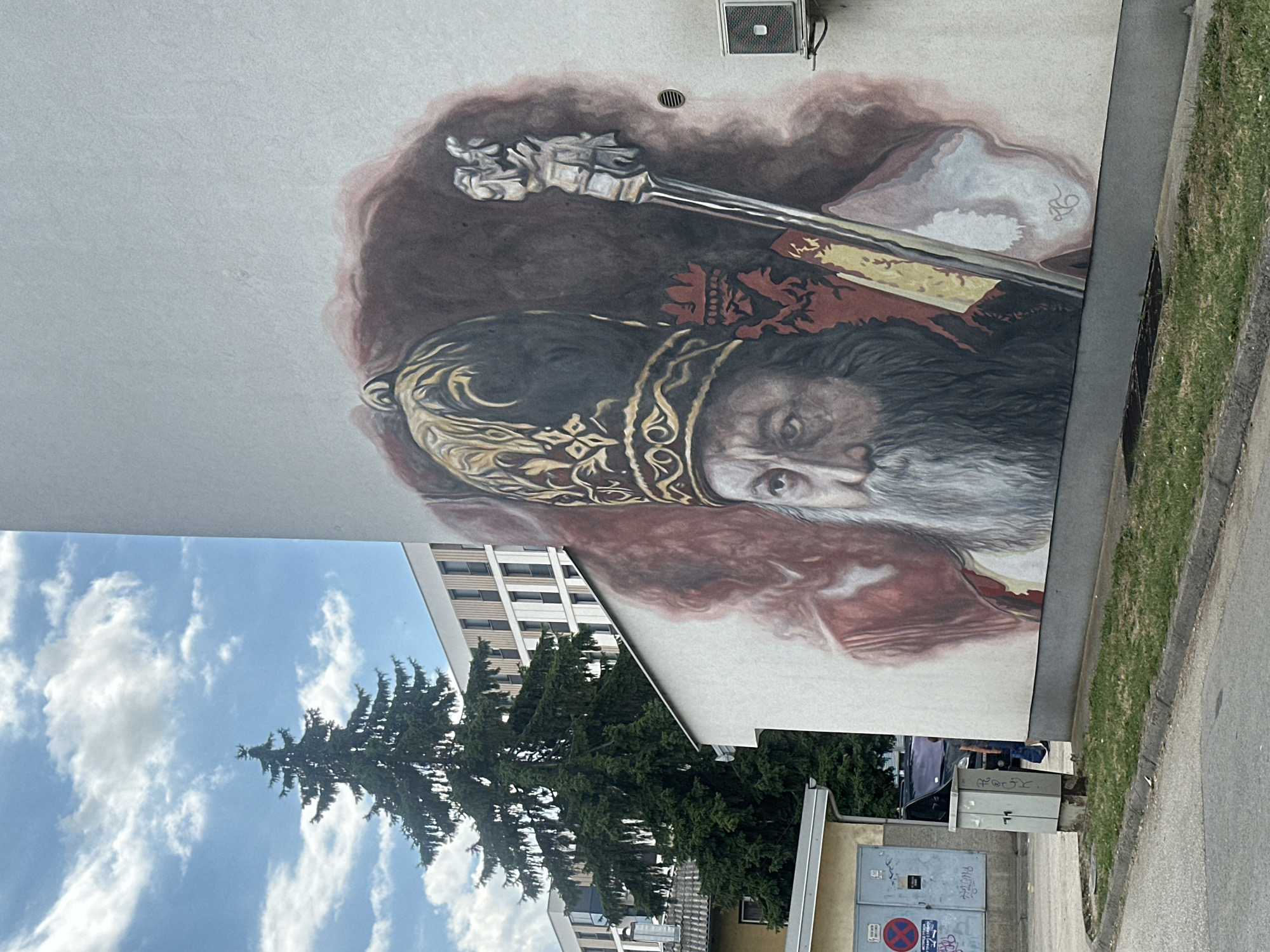 A mural of a bearded man wearing a golden, jeweled headdress and a red and gold robe is painted on the side of a white building. The man holds a silver staff in his right hand. The mural is located on the exterior wall of a building in Doboj, Bosnia and Herzegovina, near a street with a curb and a patch of grass. In the background, a tall green coniferous tree stands in front of a modern white building with many windows. A grey electrical box is positioned on the ground in front of the mural. An air conditioning unit is mounted on the wall to the right of the mural. The sky is blue with white, scattered clouds.
