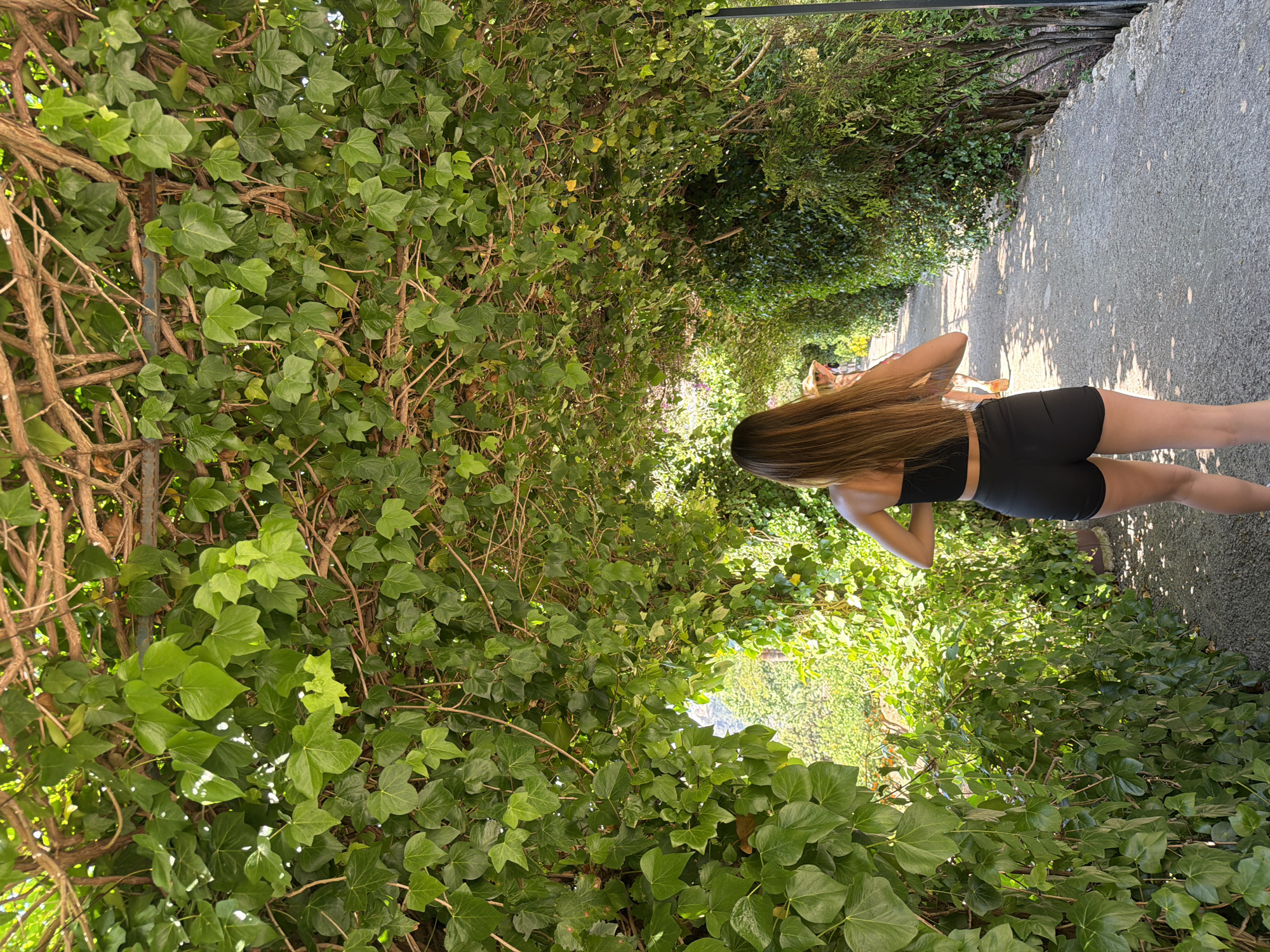 A woman in athletic wear is walking along a path lined with dense ivy foliage in Bunyola, Spain. The path is paved and leads toward the background where other people are faintly visible. The ivy forms an overhead arch.