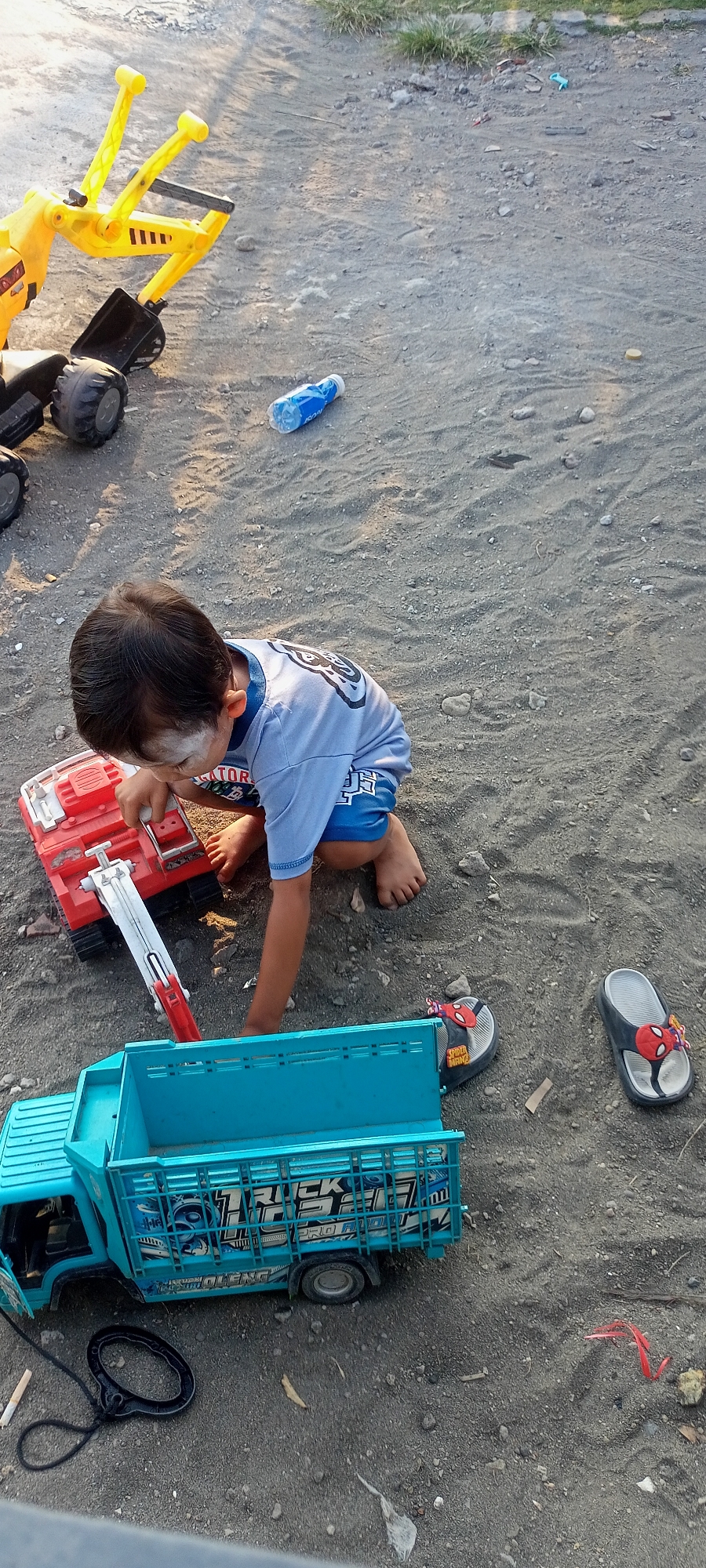 The image depicts a child playing with toys on a sandy ground in Gempolklutuk, Indonesia. The child, wearing a blue shirt and shorts, is crouched down, interacting with a red toy vehicle. In front of the child is a bright blue toy truck. Nearby, there are yellow construction toy vehicles and a bottle of water. A pair of child-sized flip-flops with a Spider-Man design are also visible on the ground. The environment is outdoors, with dry grass and soil visible in the background, suggesting a rural or undeveloped area. The lighting indicates it is daytime.