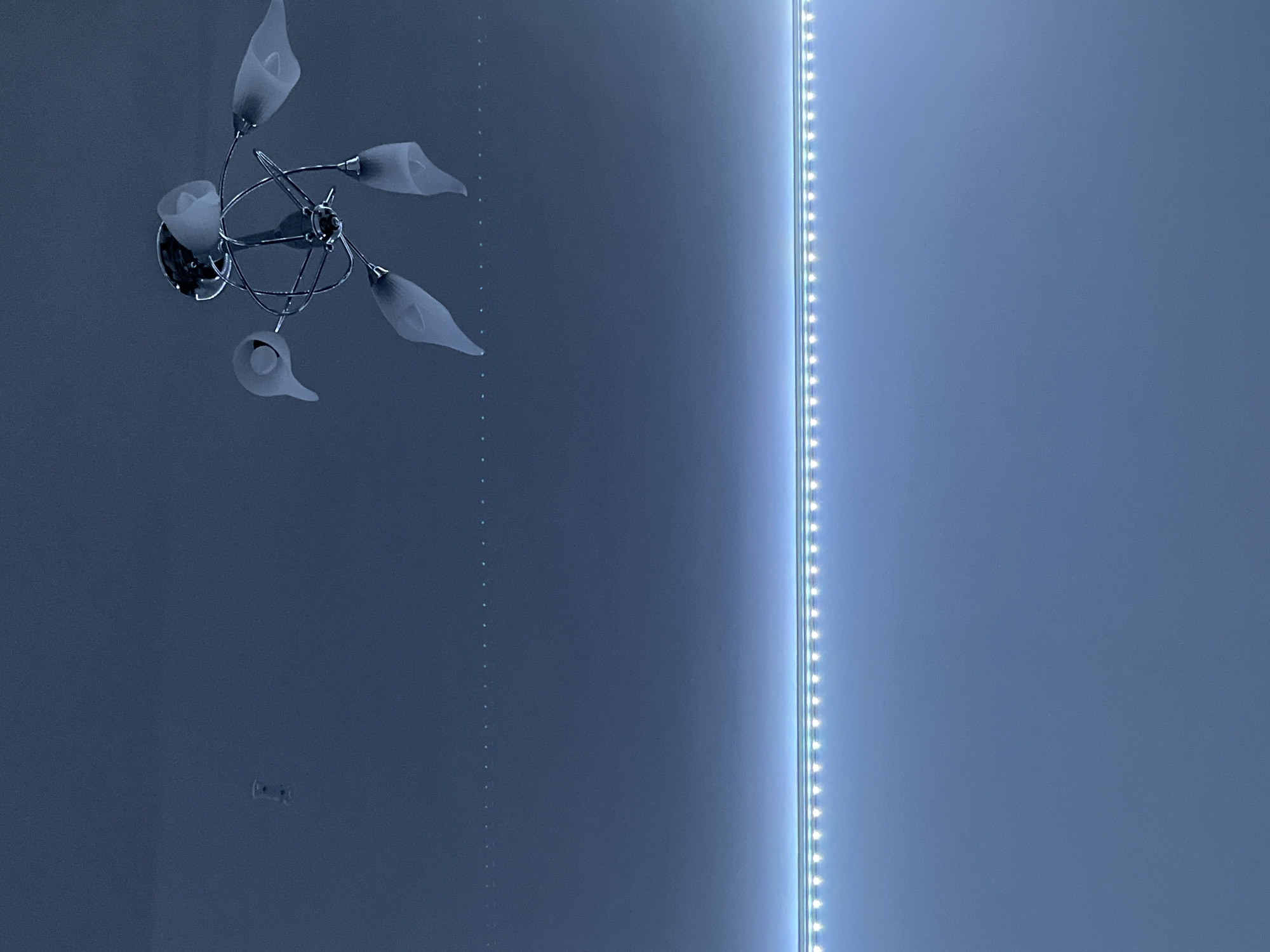 A low-angle, indoor shot shows a wall illuminated by two light sources. On the left side of the wall is a chandelier with multiple frosted glass shades arranged in a floral pattern. To the right is a vertical strip of LED lights, casting a bright, linear glow. The wall appears to be painted a muted blue. In the lower-left corner of the wall, there are visible holes where a picture, mirror, or artwork has been removed. There is no indication of location in Saint Petersburg, Russia.