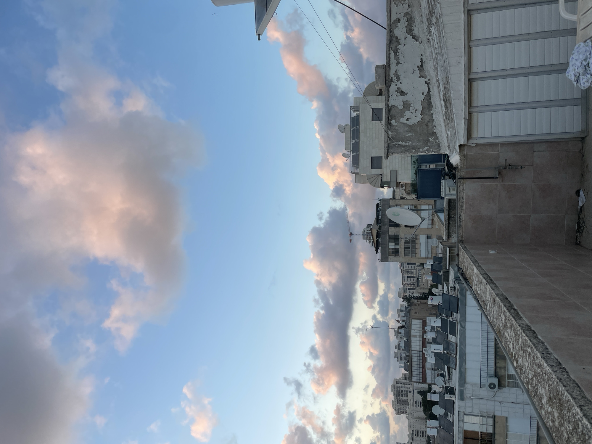 The image shows a view from a rooftop in Petah Tikva, Israel, at what appears to be dusk. The sky is filled with clouds that are illuminated with a pink hue from the setting sun. Buildings of varying heights are visible in the distance, and several buildings have satellite dishes on their roofs. In the foreground, there is a concrete ledge. A window or doorway with horizontal panels can be seen to the right of the ledge.