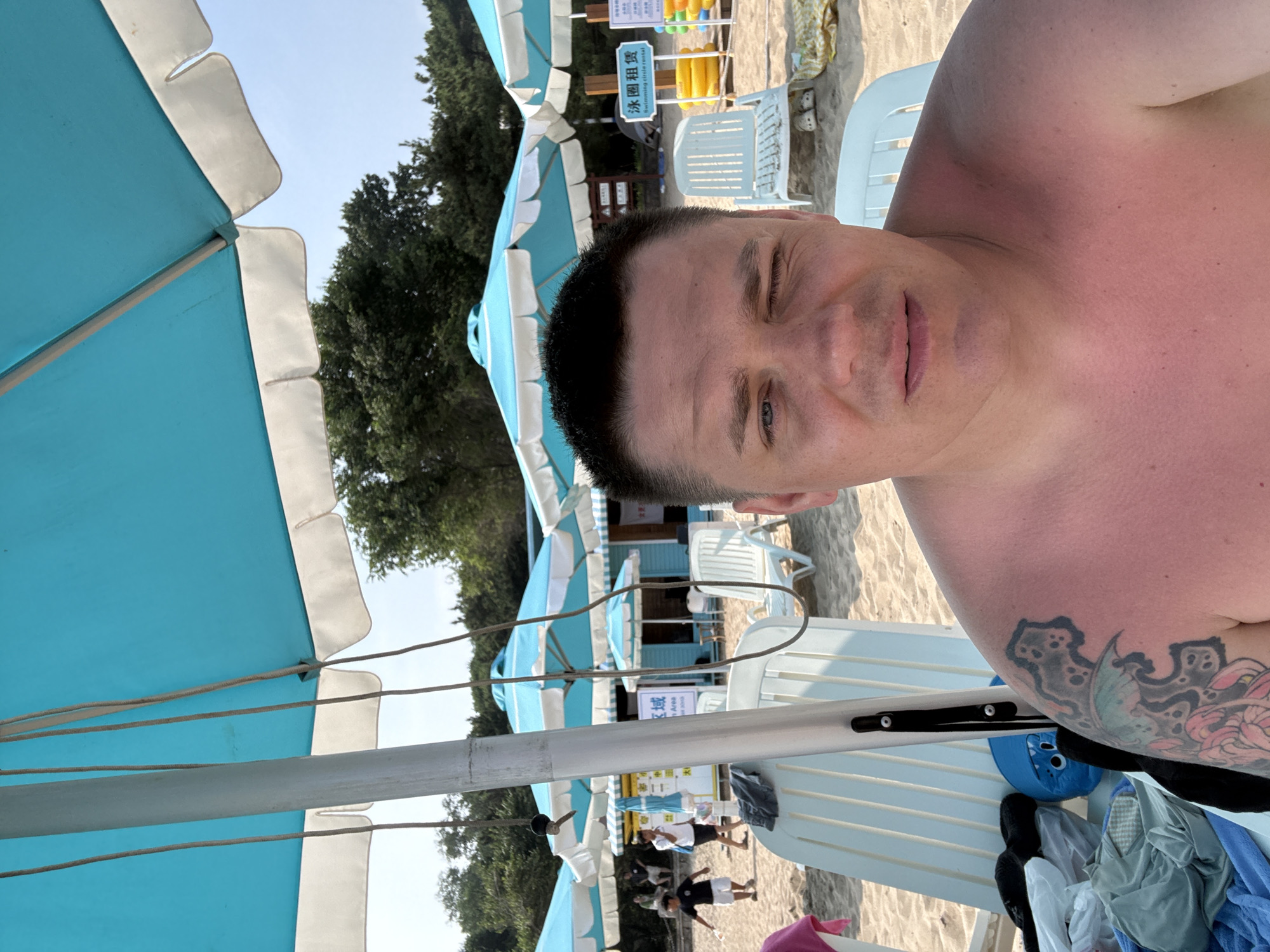 The image shows a man taking a selfie on a sandy beach in Qinhuangdao, China, likely during daytime. He is shirtless, with visible sunburn on his shoulders and chest, and has a tattoo on his left arm. He is winking. In the background, there are several blue and white beach umbrellas and white lounge chairs. Behind the chairs, there is a building with signage in Chinese characters, and further back, a line of trees. Several people are visible in the distance, some walking and others sitting. Yellow and orange swimming rings are stacked near a sign that reads "Swimming circle rental" in English. The overall scene suggests a popular beach resort area.