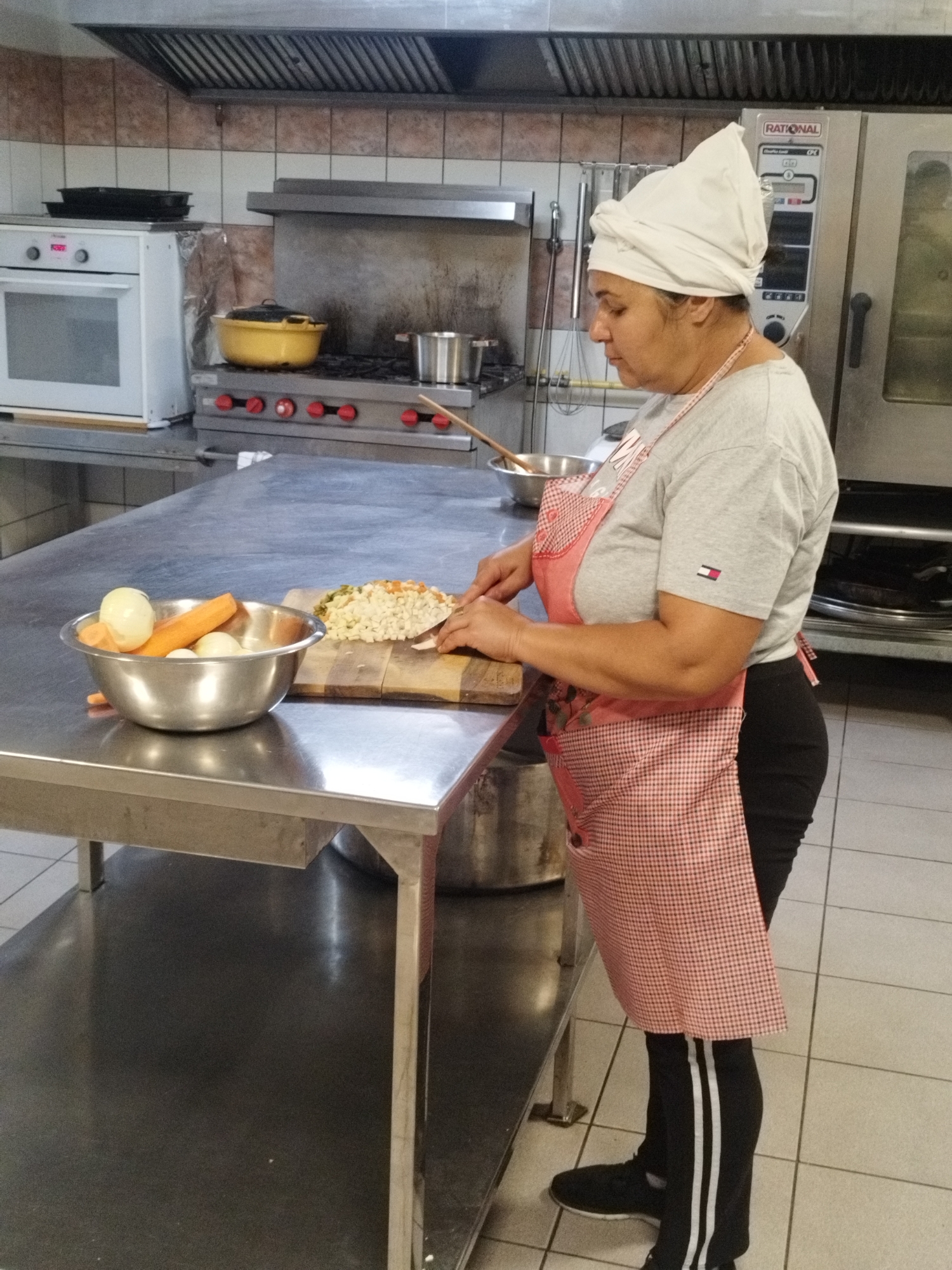 In a commercial kitchen, a woman wearing a white head covering and a red and white checkered apron stands at a stainless steel table, preparing food. She is in the process of chopping vegetables, with a cutting board and a pile of chopped vegetables in front of her. A bowl containing whole onions and carrots is also on the table. In the background, commercial kitchen equipment is visible, including ovens and stovetops. The environment appears to be a professional kitchen setting, likely in Bucharest, Romania, given the context.