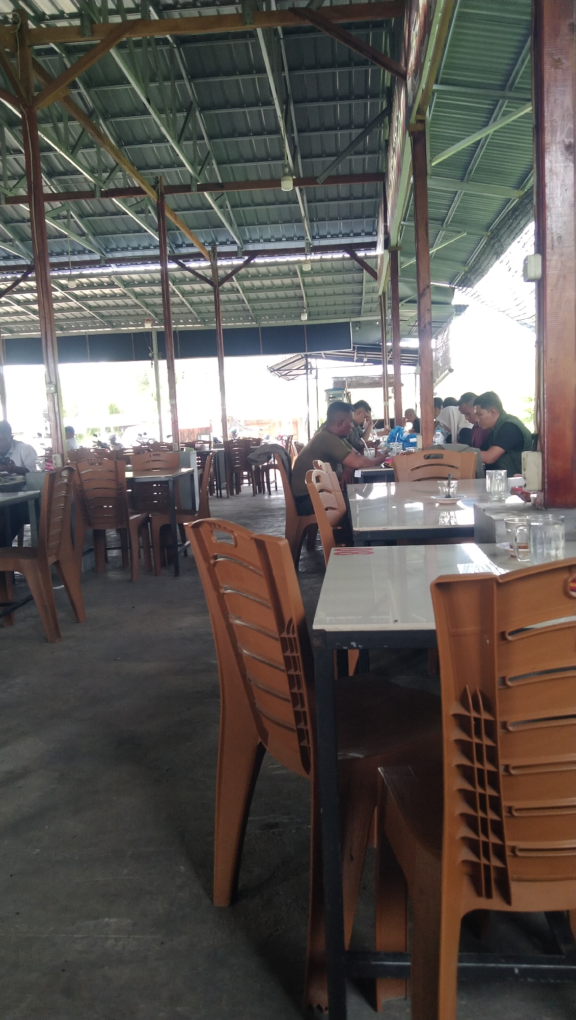 The image depicts an outdoor seating area of a restaurant or food court in Keude Trienggadeng, Indonesia. The scene is covered by a metal roof supported by wooden and metal beams, with some natural light filtering in. Numerous tables and brown plastic chairs are arranged throughout the space. In the background, several people are seated at tables, some eating or conversing. The flooring appears to be concrete. The overall impression is of a casual dining establishment.