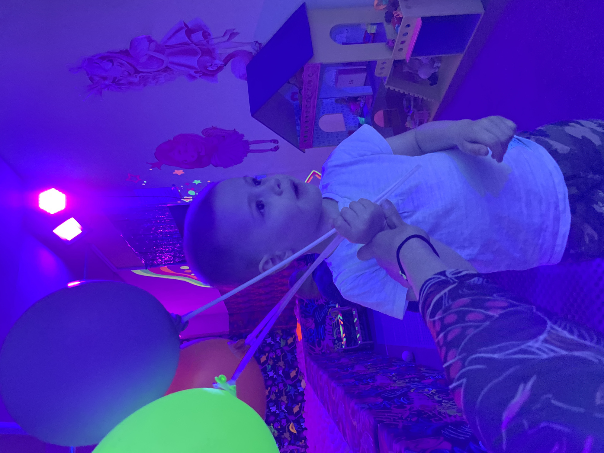 In a room illuminated by ultraviolet or blacklight, a young child stands beside a table, surrounded by brightly colored balloons. The room, possibly a playroom or event space in an Unknown city, features a dollhouse, princess decorations on the ceiling, and other toys. A person's arm is visible, perhaps assisting the child with the balloons.