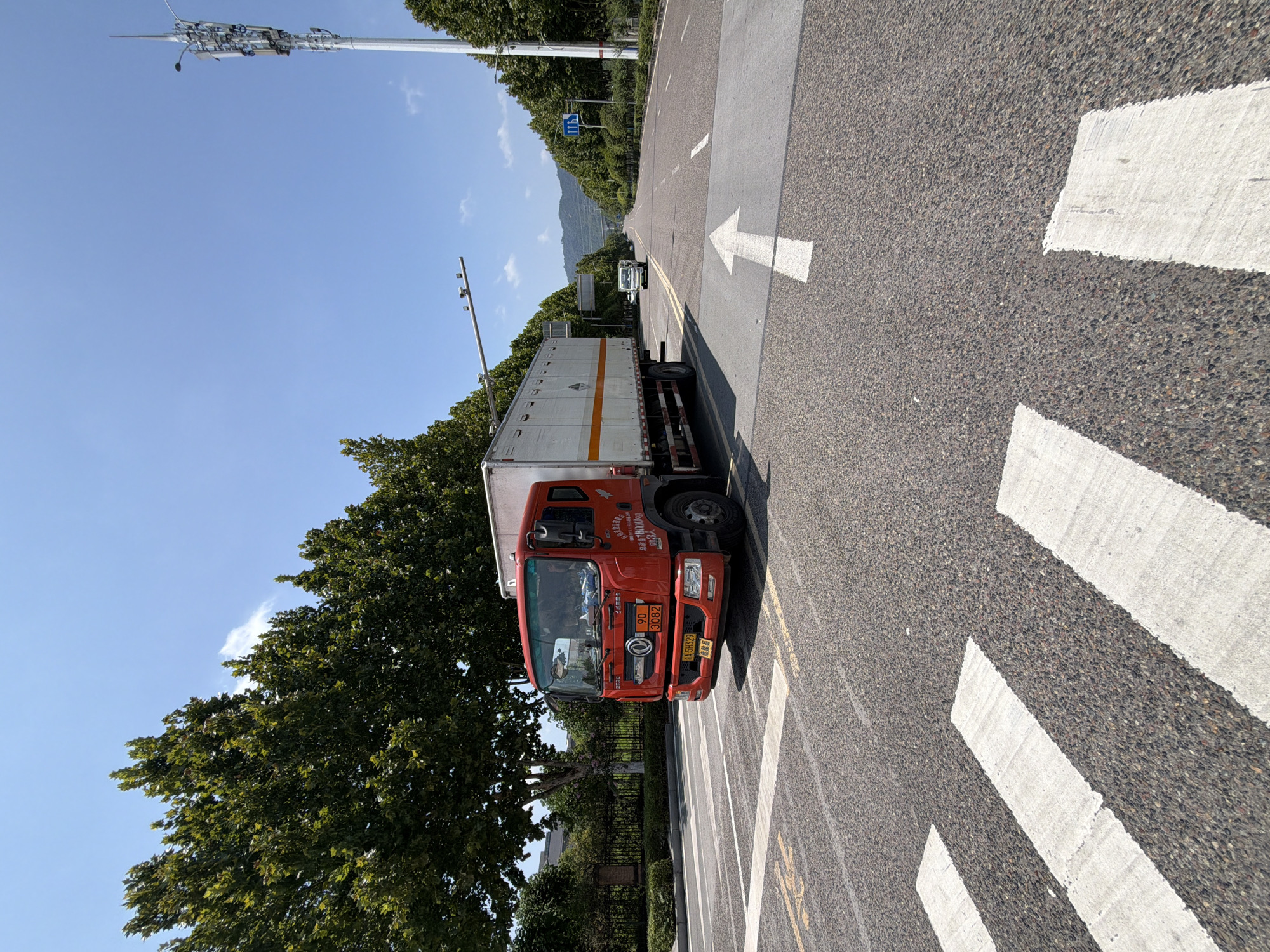 The image shows a bright, sunny day in Jiangjin, China, with a red truck stopped at a pedestrian crossing on a wide asphalt road. The truck, a medium-duty commercial vehicle, has a white cargo box with an orange stripe. It is facing towards the right of the frame, with its front visible. The road is marked with white lines for the pedestrian crossing and a white arrow indicating a forward direction. Trees line both sides of the road, and in the distance, there are hills under a clear blue sky. A tall telecommunications tower is visible on the right side of the road. A white car is seen further down the road, also heading away from the viewer. The scene appears to be in an urban or suburban area.