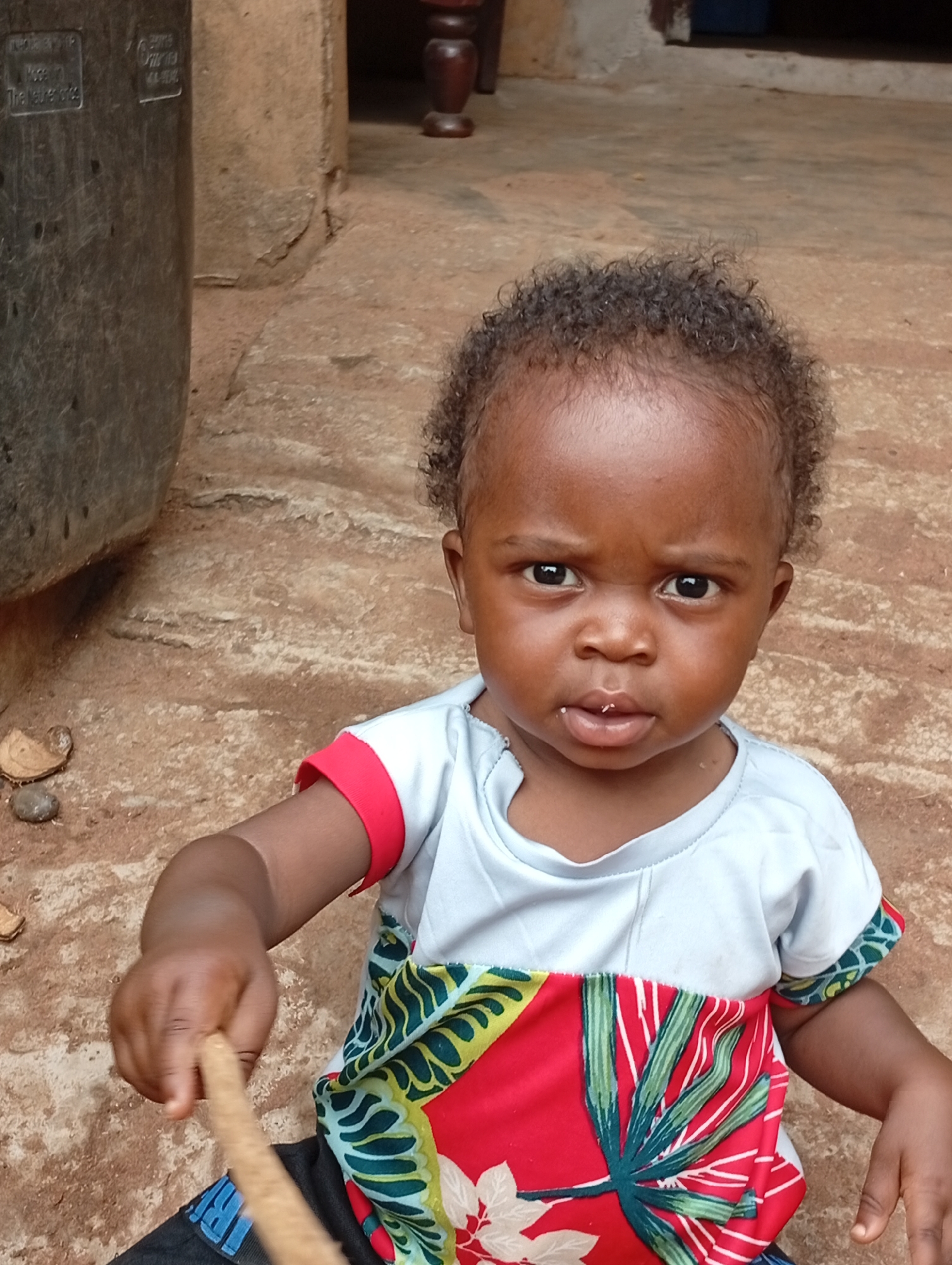 A young child with curly hair and dark skin is pictured sitting on a dirt ground in what appears to be a rural setting in Adda, Nigeria. The child is wearing a light blue t-shirt with red trim on the sleeves and a colorful pattern on the front featuring red, green, and white floral designs. The child is holding a light brown stick in their right hand, with their left arm resting on their lap. The child's expression is serious, with wide-open eyes and slightly parted lips. In the background, a large dark grey cylindrical object, possibly a bin or container, is visible on the left side of the image. Behind the child, the ground appears to be made of packed dirt with some scattered debris, including small stones and a piece of what looks like a nut shell. The overall environment suggests an outdoor, possibly domestic, setting.